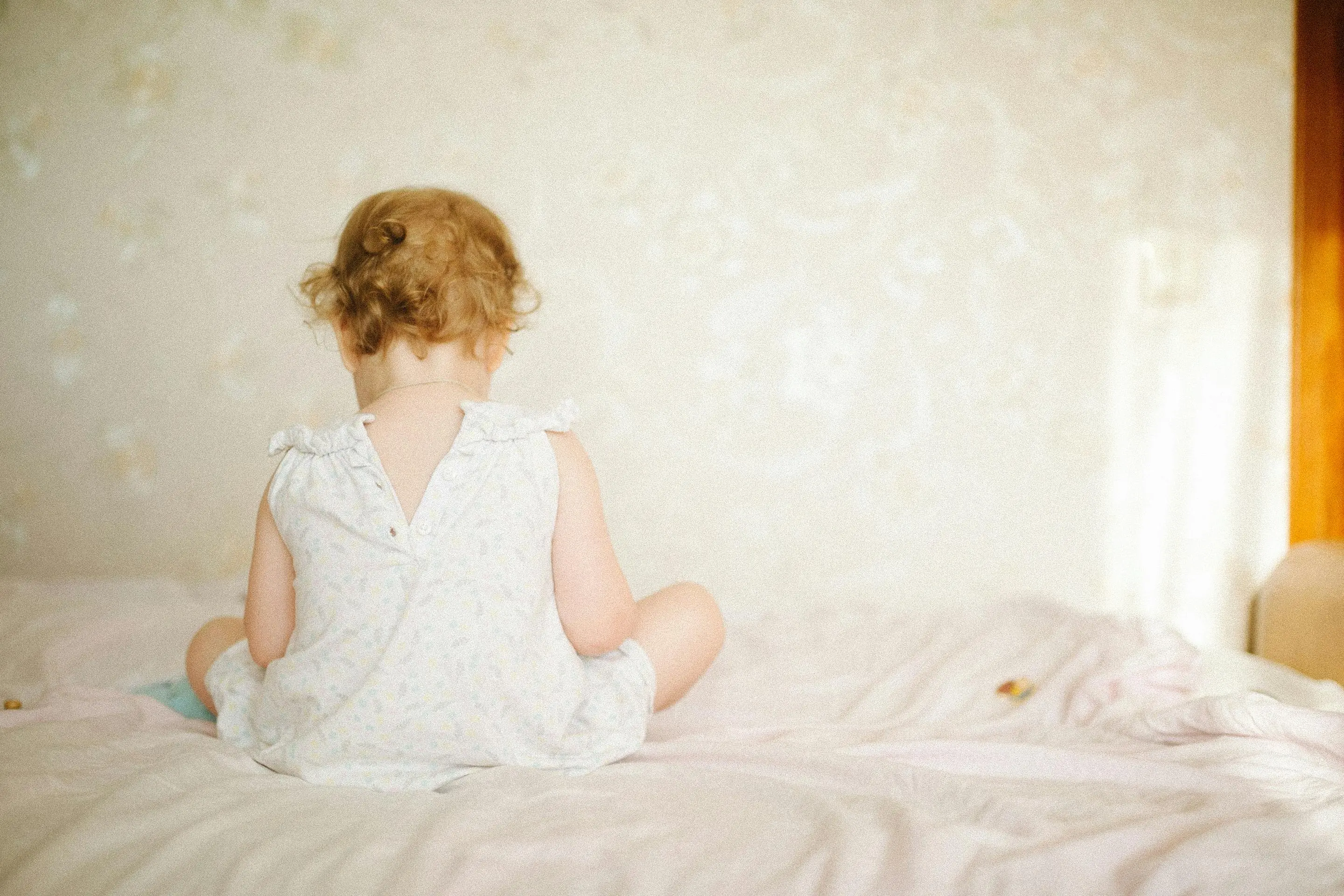 baby bed bugs A cute baby with curly hair sitting on a bed in soft natural light, back view.