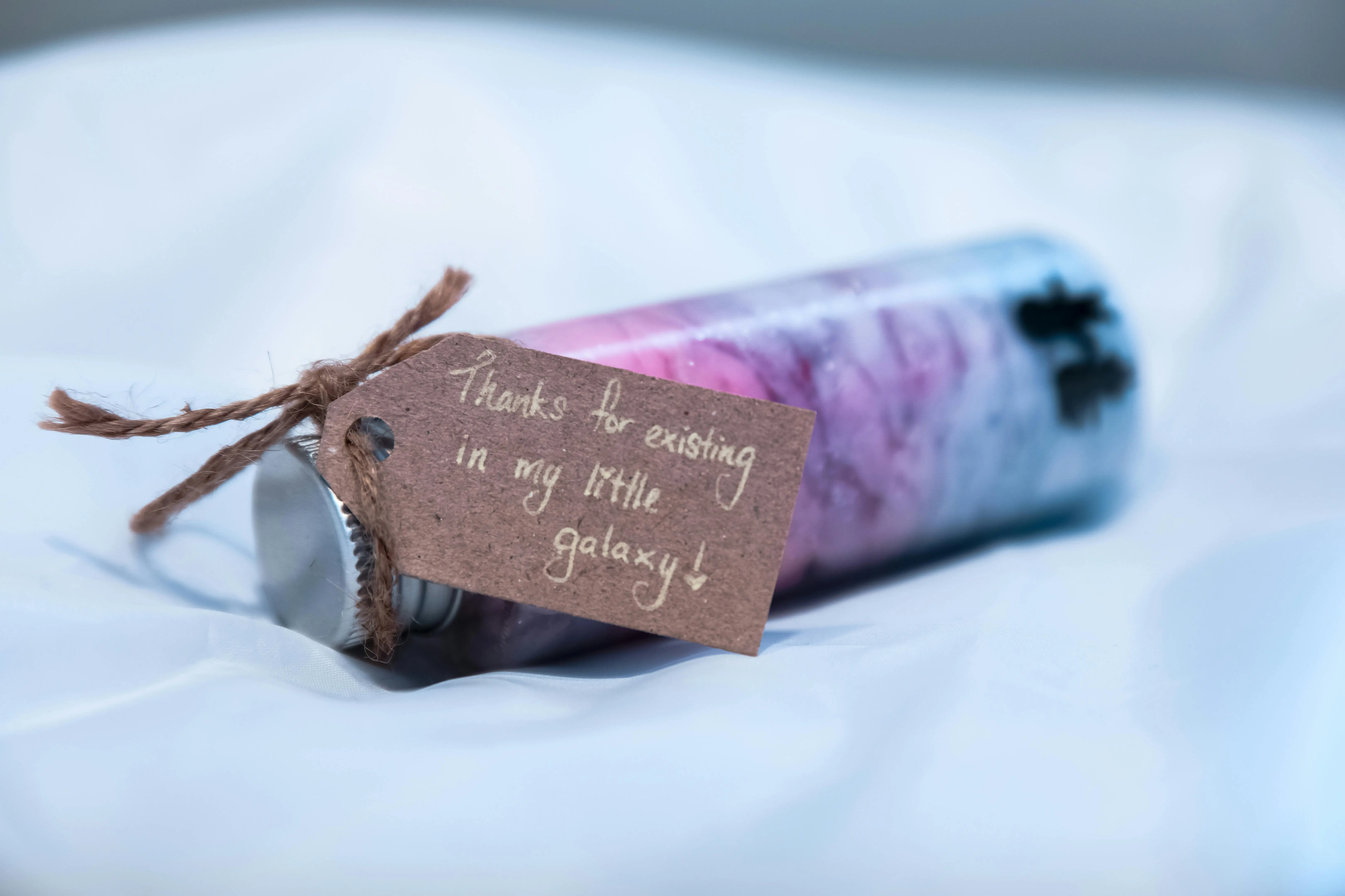 inexpensive team gifts A candle with purple hues and a heartfelt message on a kraft tag, saying 'Thanks for existing in my little galaxy'.