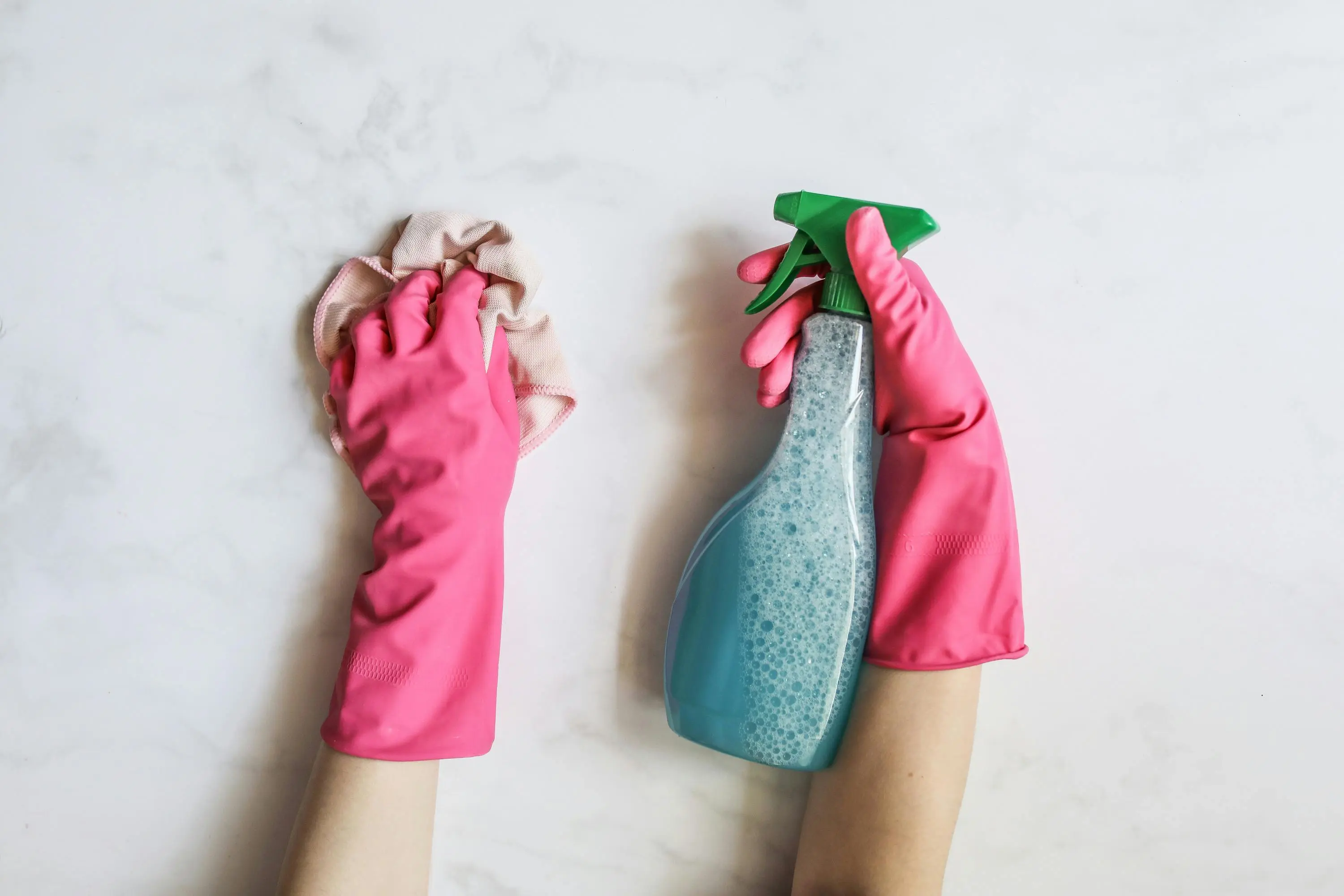 how to get mildew smell out of clothes A pair of hands in pink gloves are cleaning with a spray bottle on a white background.