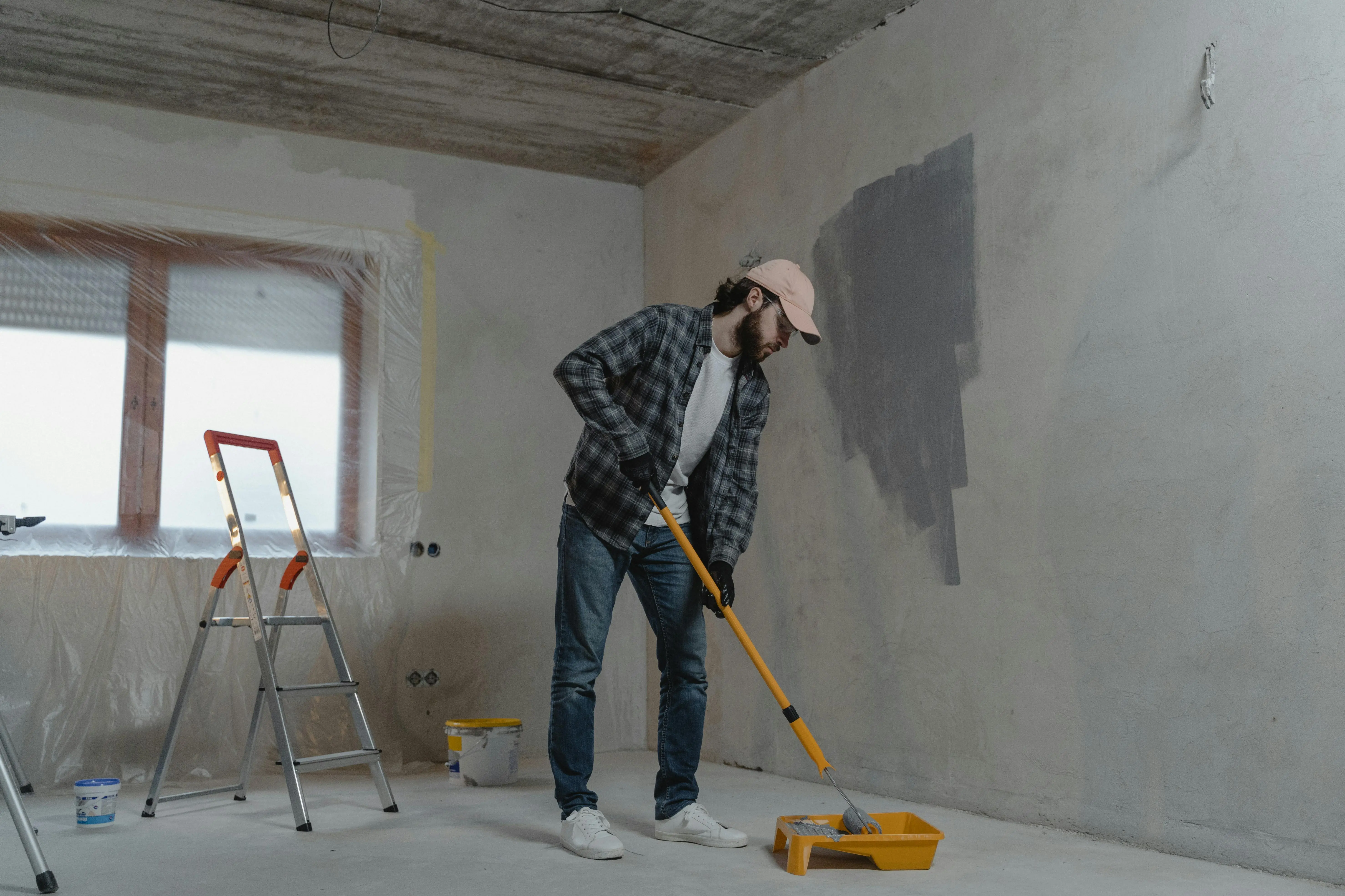 grey paint A man painting a room with a roller, using a stepladder, wearing casual attire. Renovation scene.