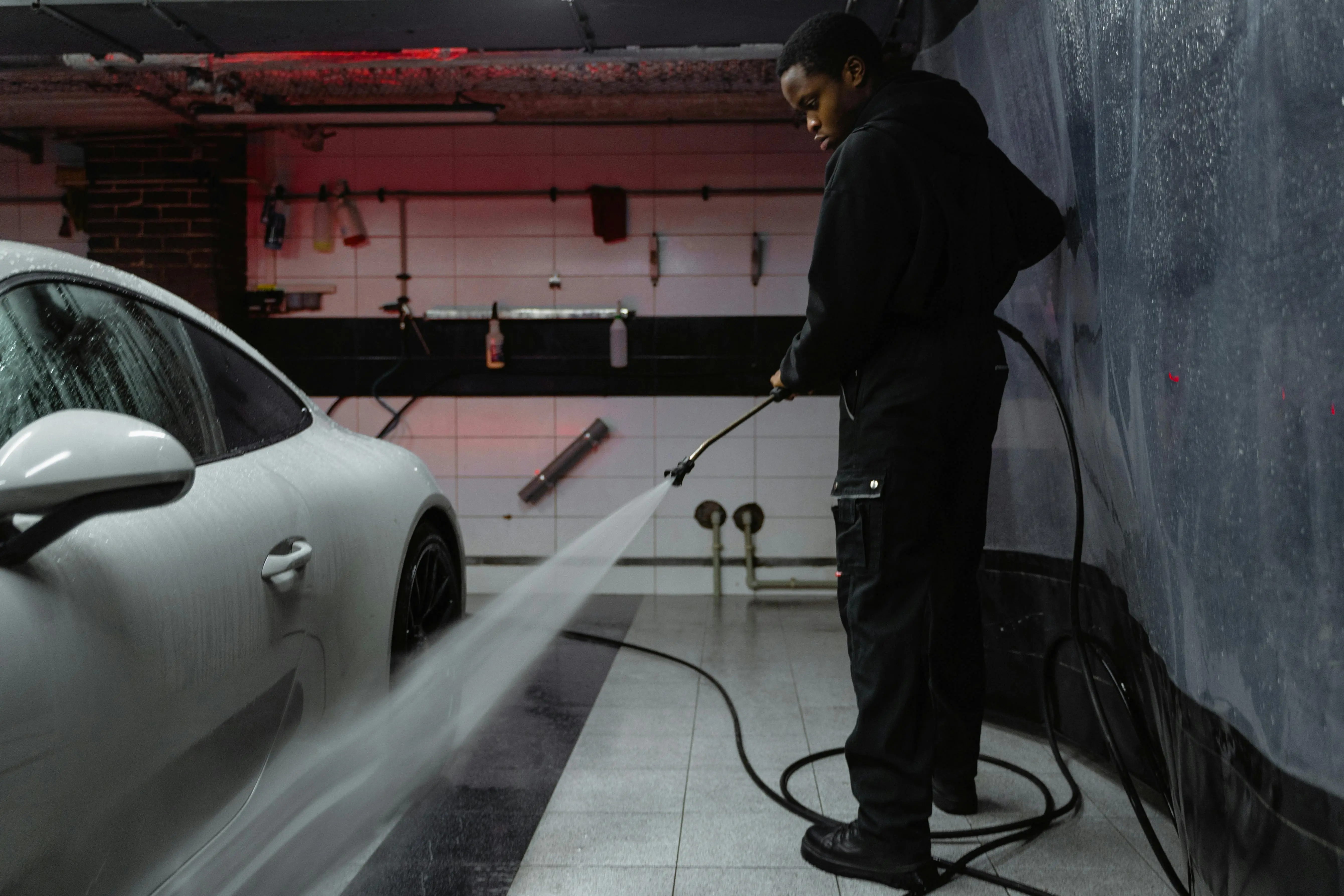 how to wash silk African American man cleaning a white car with a pressure hose in an indoor garage setting.