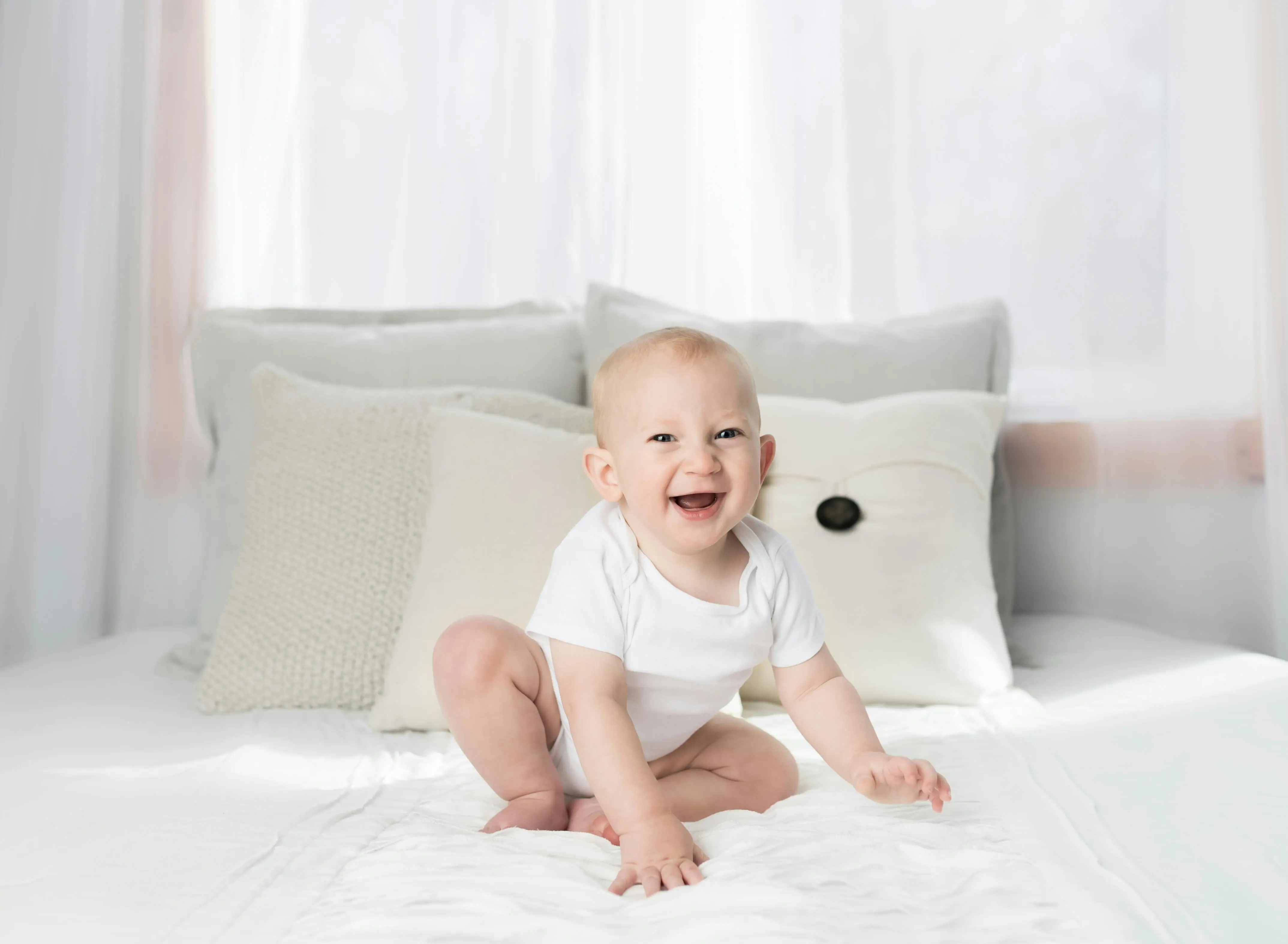 fragrance free laundry detergent Cute baby sitting and smiling on a white bed in a bright, airy bedroom.