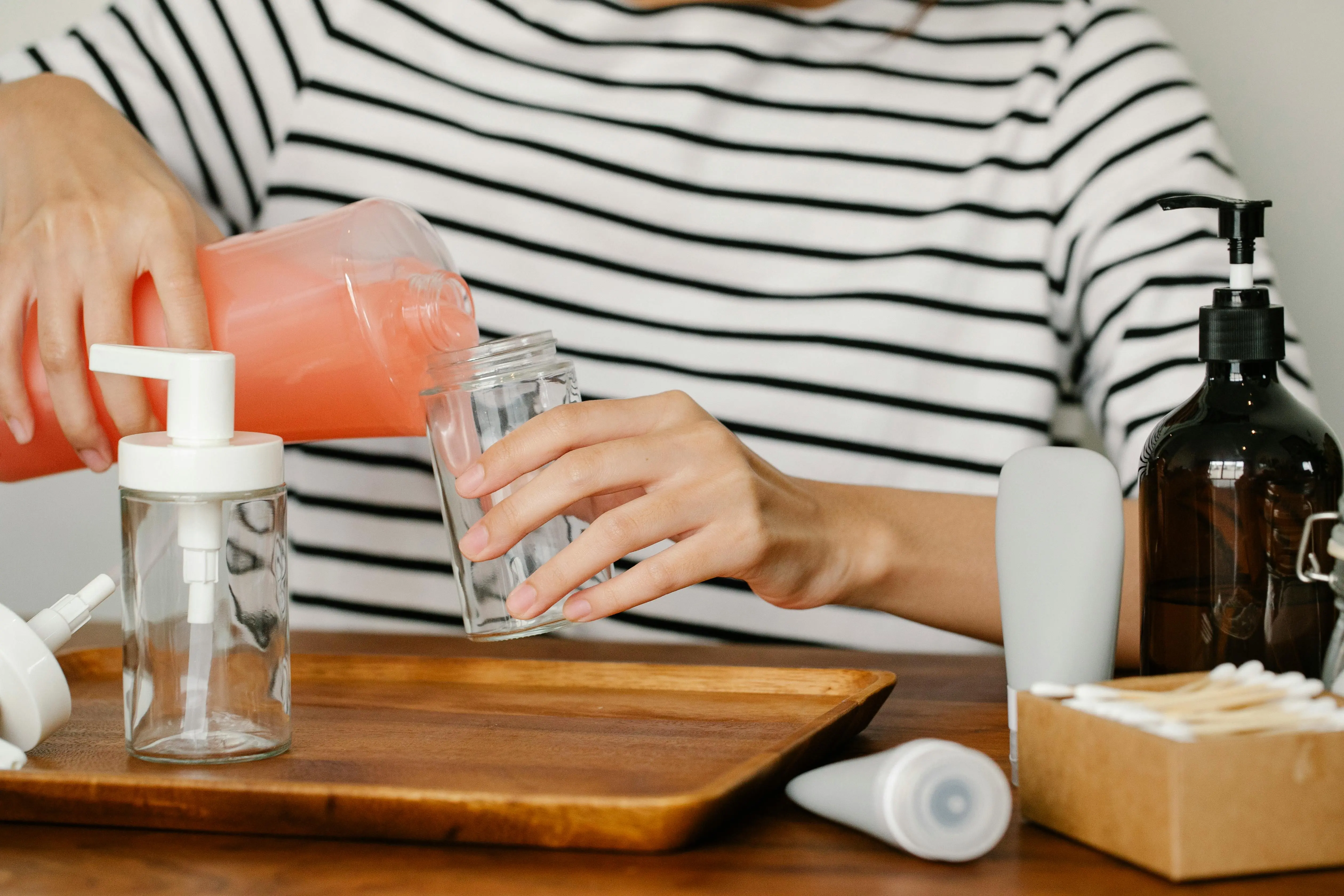 fragrance free laundry detergent Cropped view of woman pouring pink liquid into glass dispenser bottle on wooden tray.