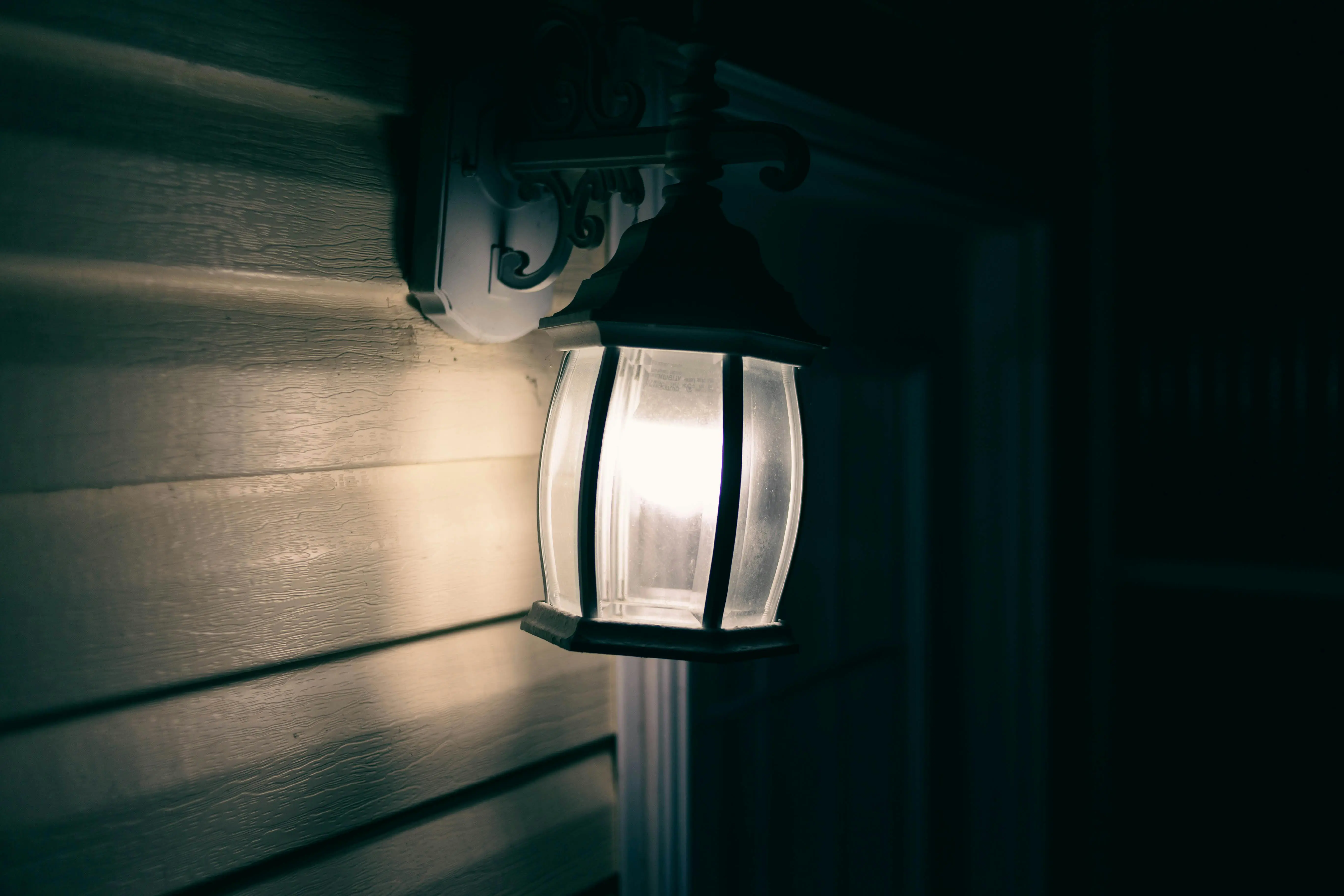 exterior wall sconce lights A close-up of an illuminated outdoor wall lamp casting a warm glow on wooden siding.
