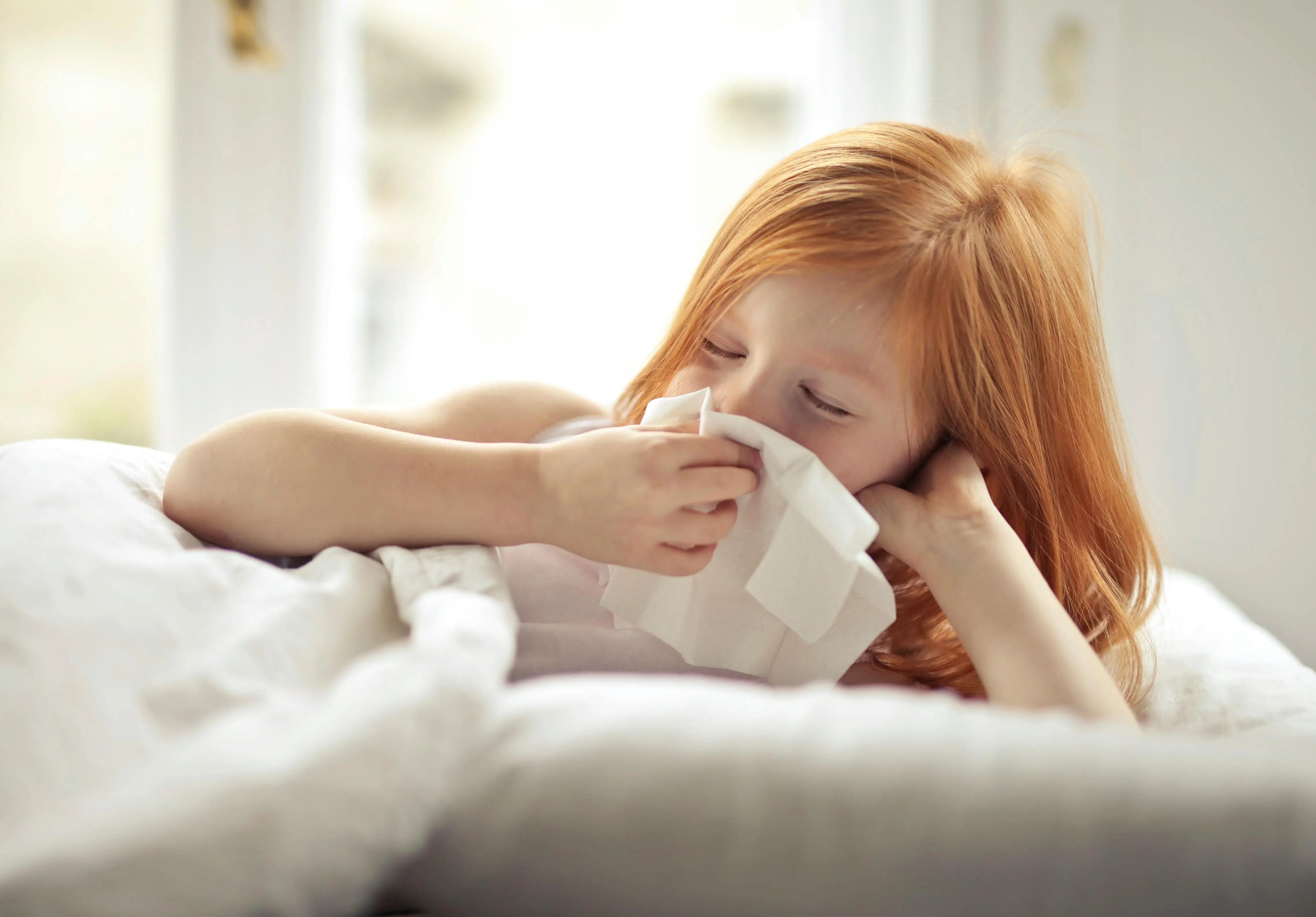 clean steam Small sick redhead kid with eyes closed cleaning nose with white tissue paper while lying alone in bed in light modern bedroom in daytime