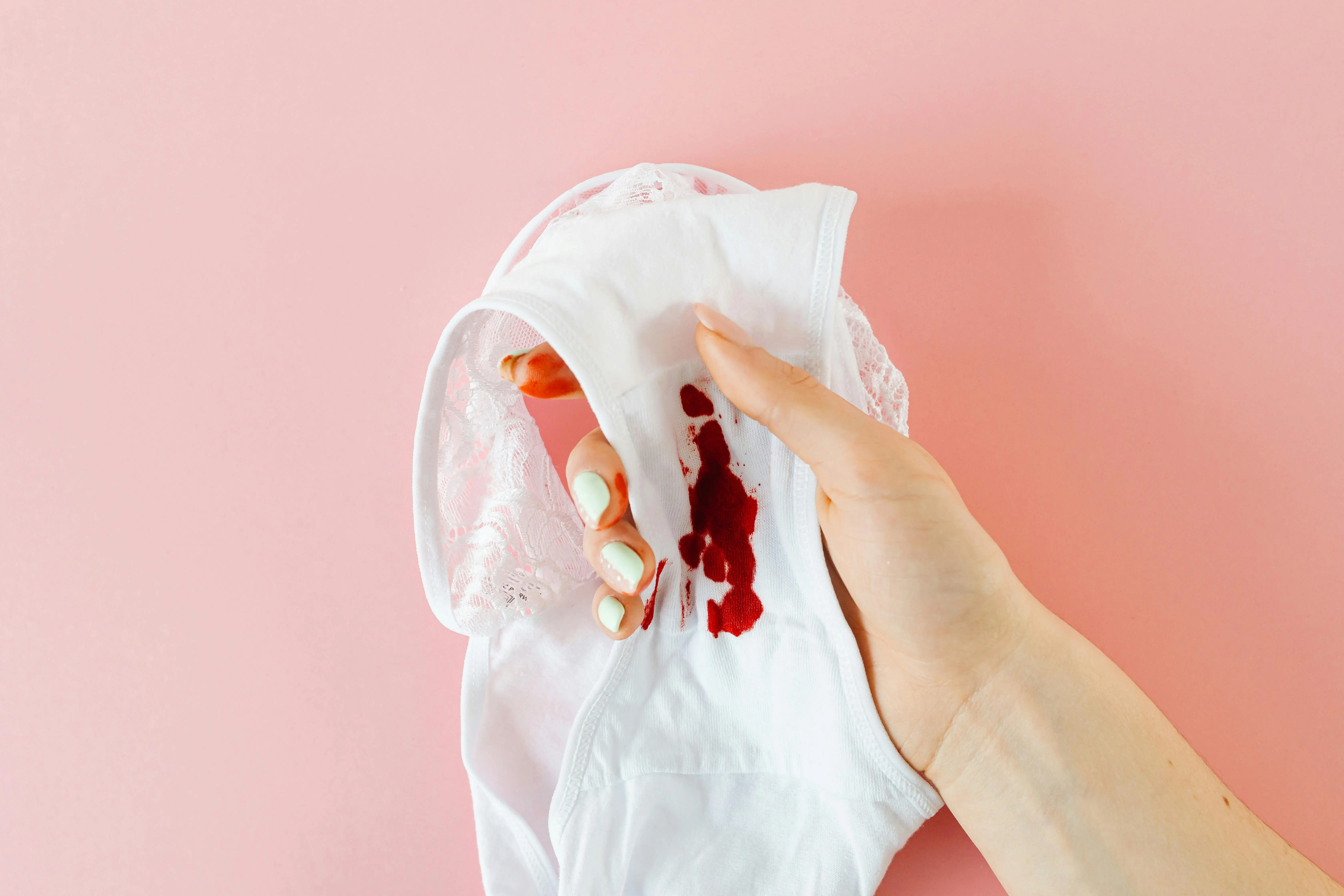 how to get rid of bleach stains Close-up of a hand holding white underwear with menstrual stain against pink background, highlighting health awareness.
