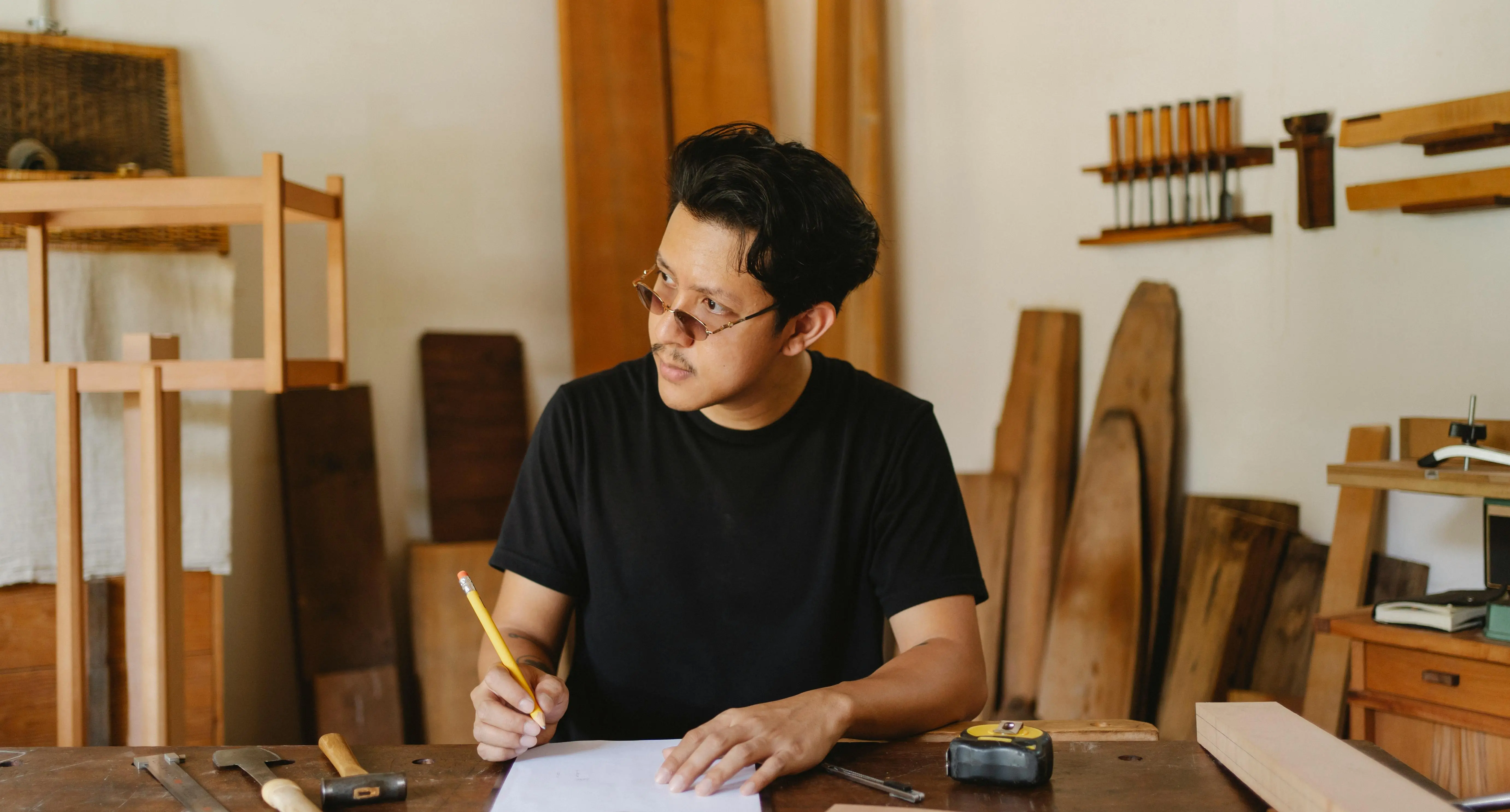 carpenter with tools Pensive ethnic male designer in casual clothes drawing sketches of new woodwork object while working in studio