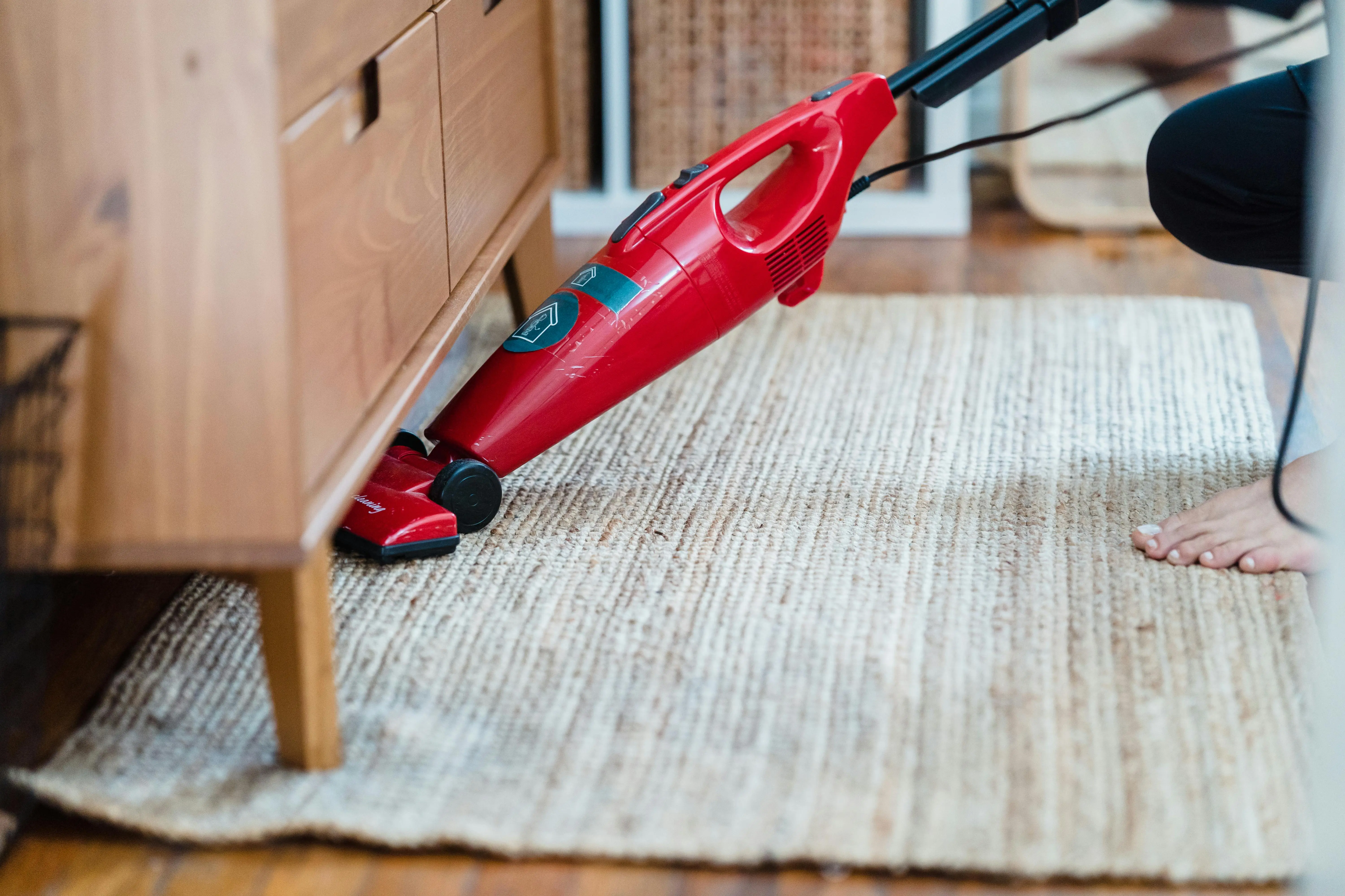 how to get rid of bleach stains Side view of a person using a red vacuum under furniture on a rug for effective cleaning.