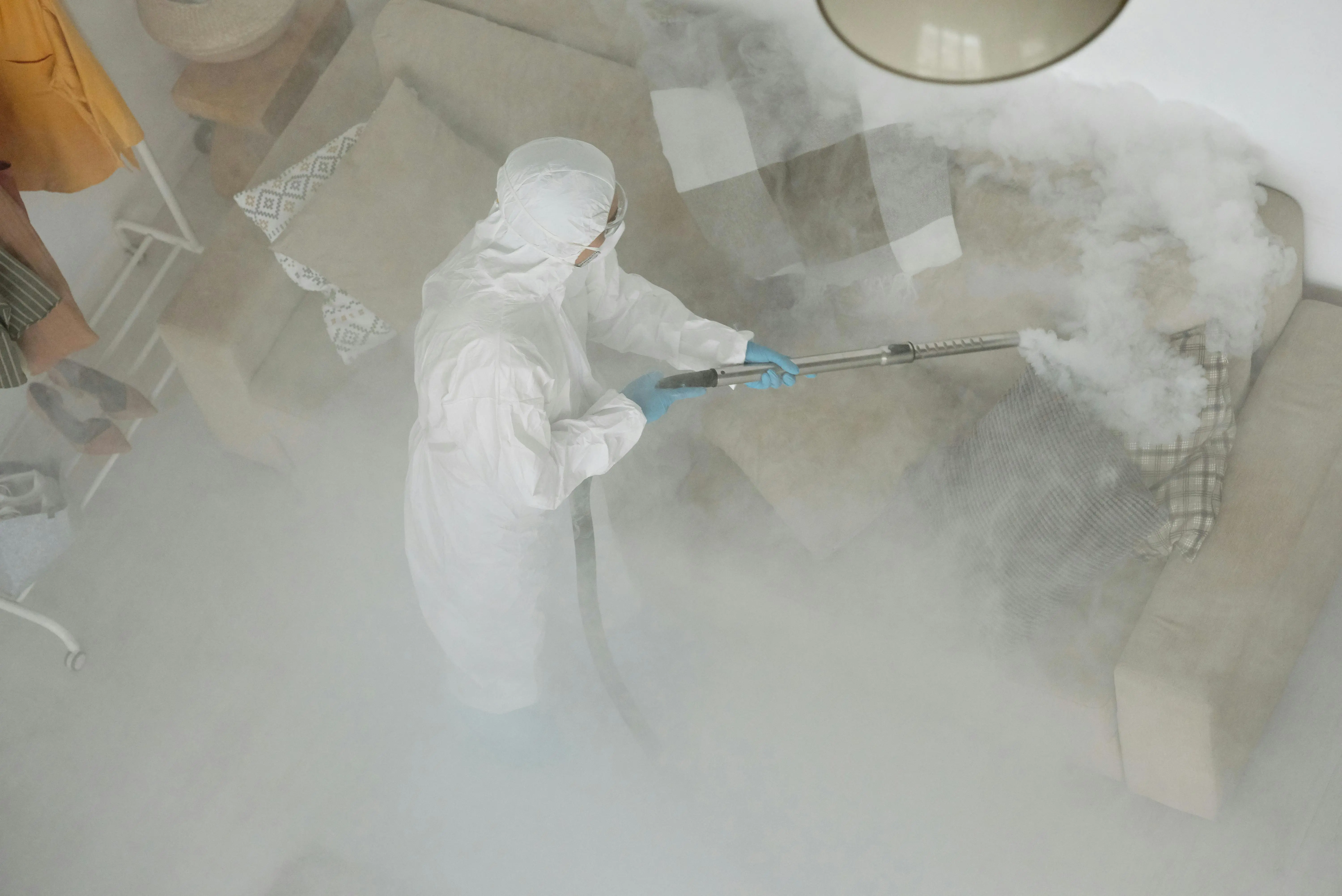 how to clean upholstered furniture Technician in protective gear sanitizing a living room with smoke fumigation.