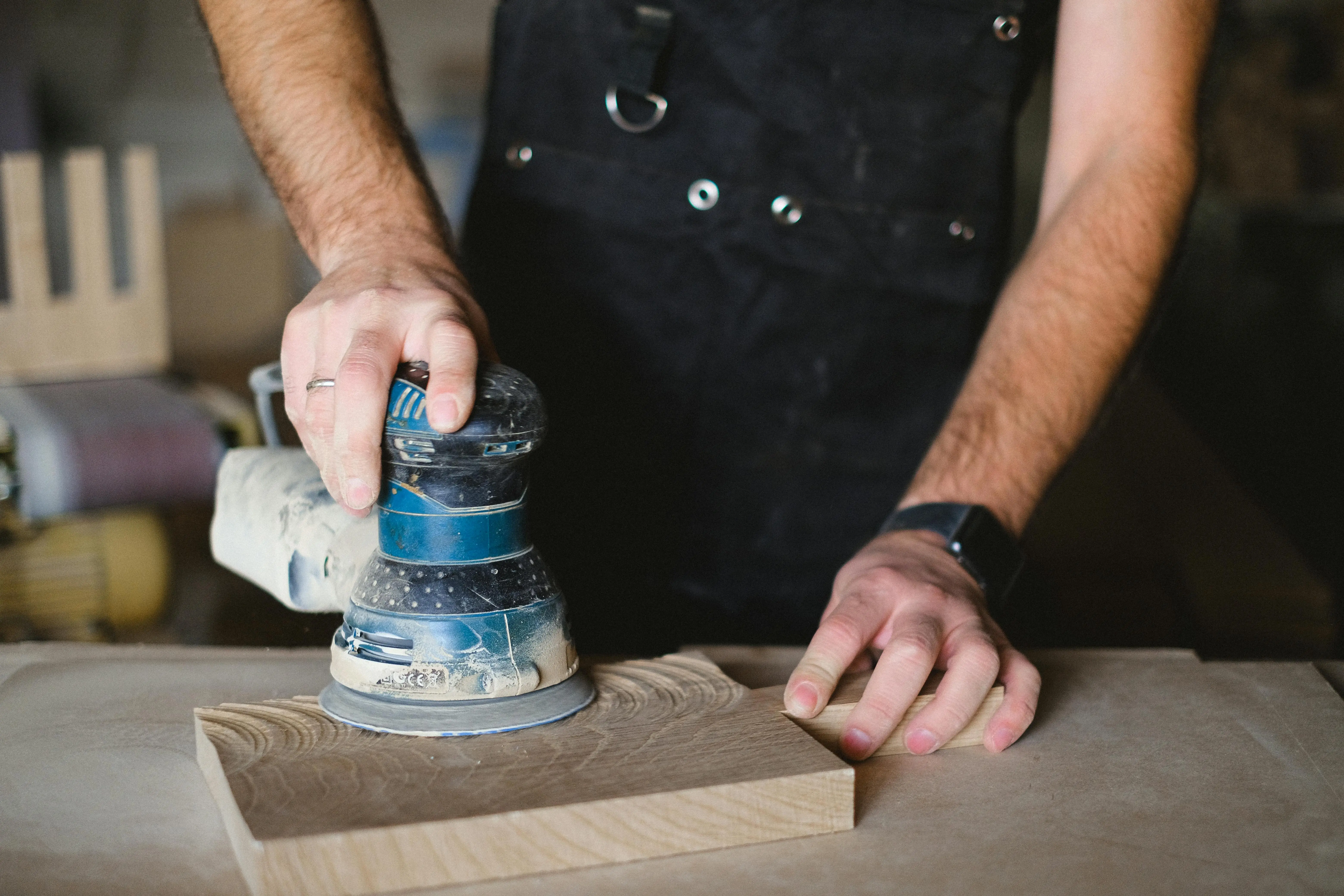 diy wood floor refinishing Unrecognizable male carpenter using random orbital sander while working with wooden detail at workbench in professional workshop on blurred background