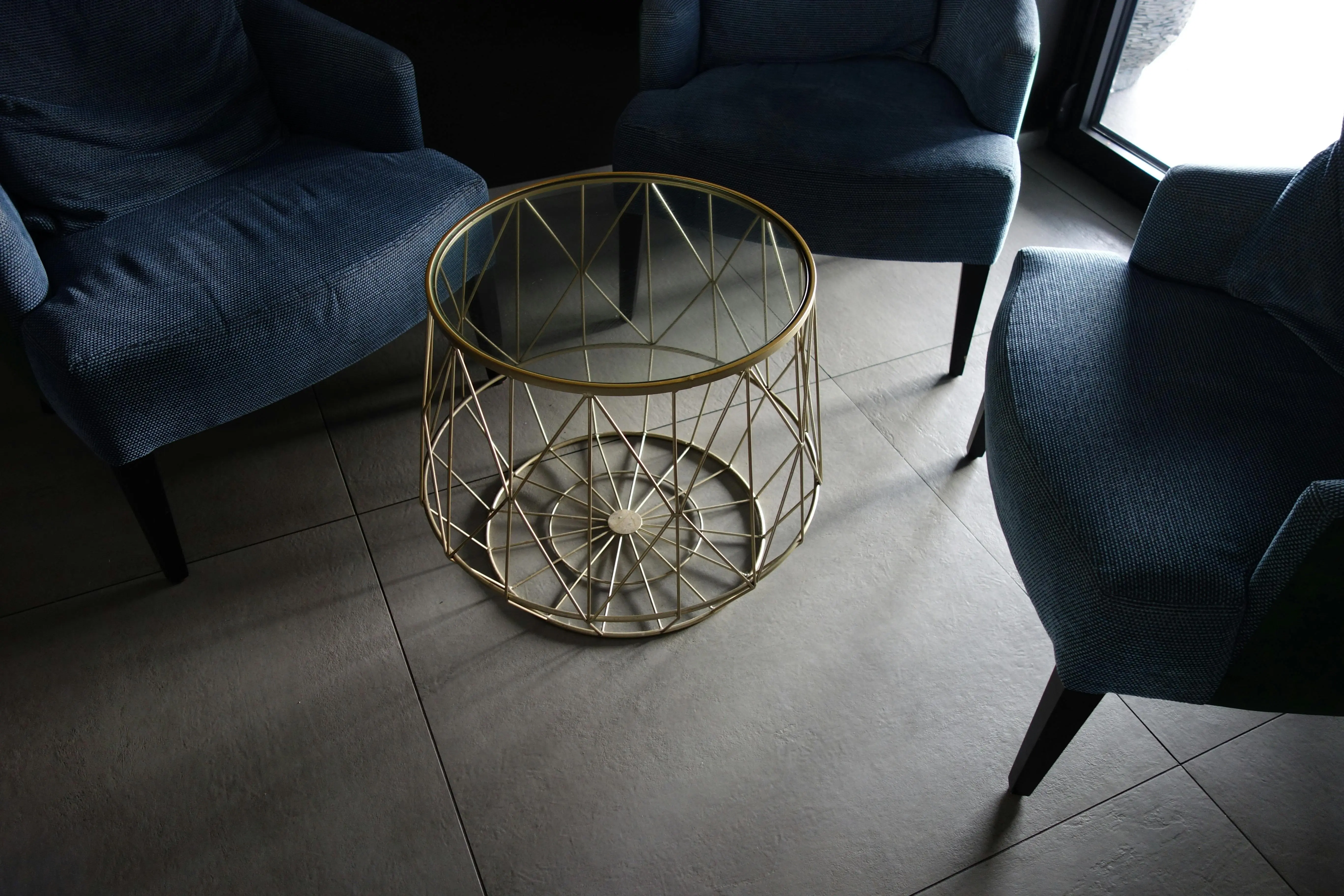 what might be placed in a secondary furniture Stylish indoor lounge featuring blue armchairs around a glass-top metal table on tiled flooring.