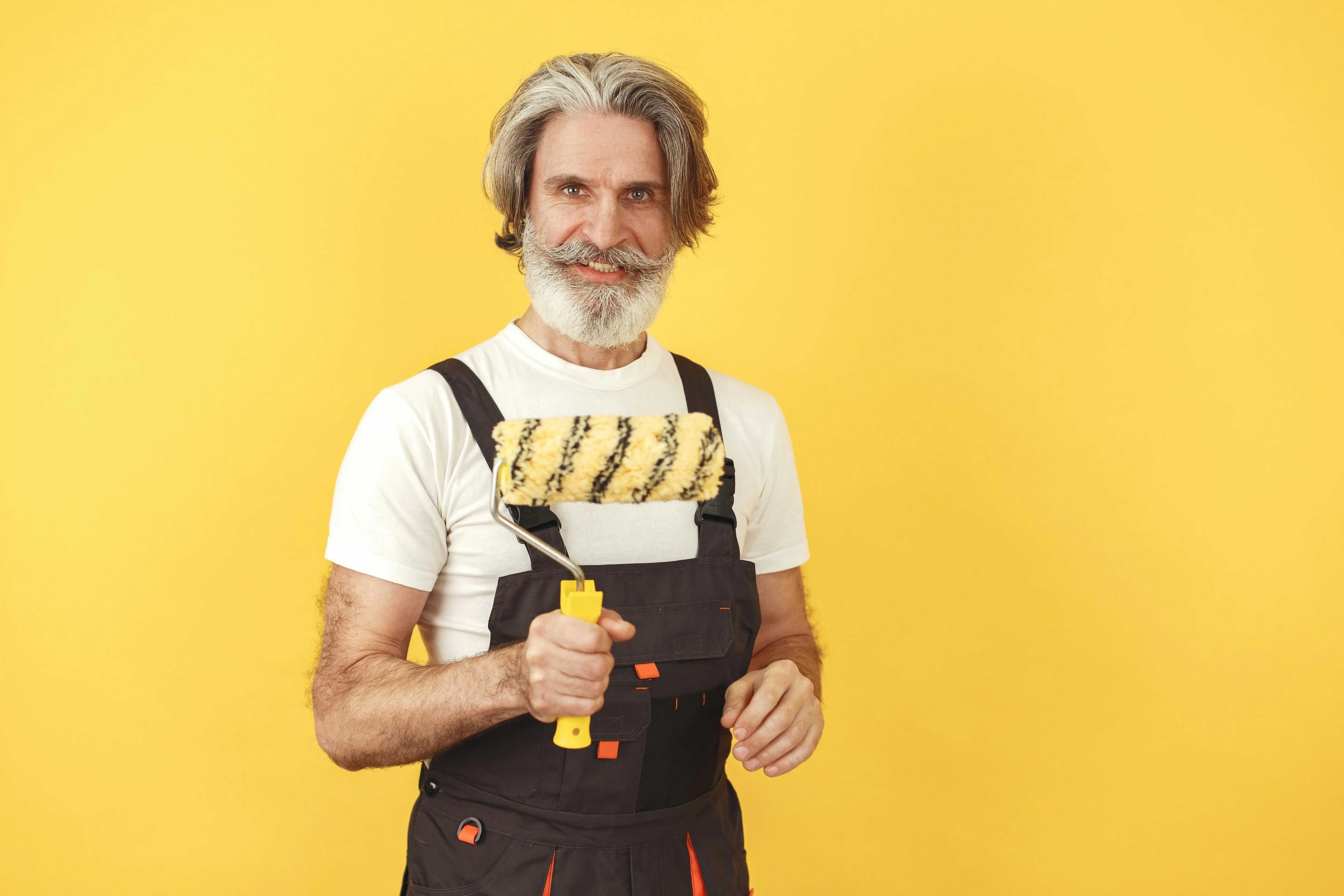 how much does a handyman charge to paint a room Senior man in overalls holding a paint roller against a vibrant yellow background.
