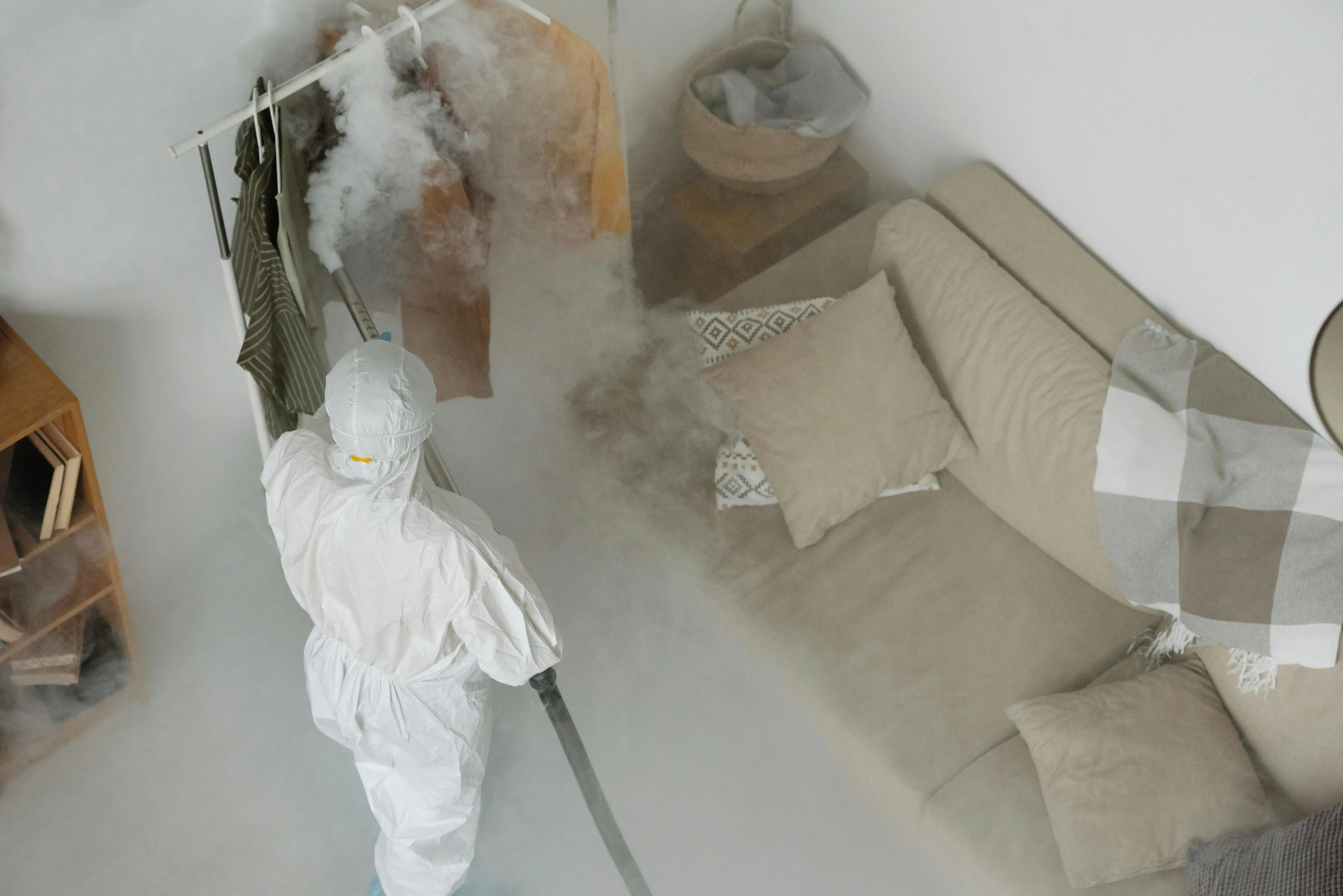 how to get rid of mildew smell in clothes A worker in protective gear performs fumigation in a living room, using smoke to sanitize surfaces.