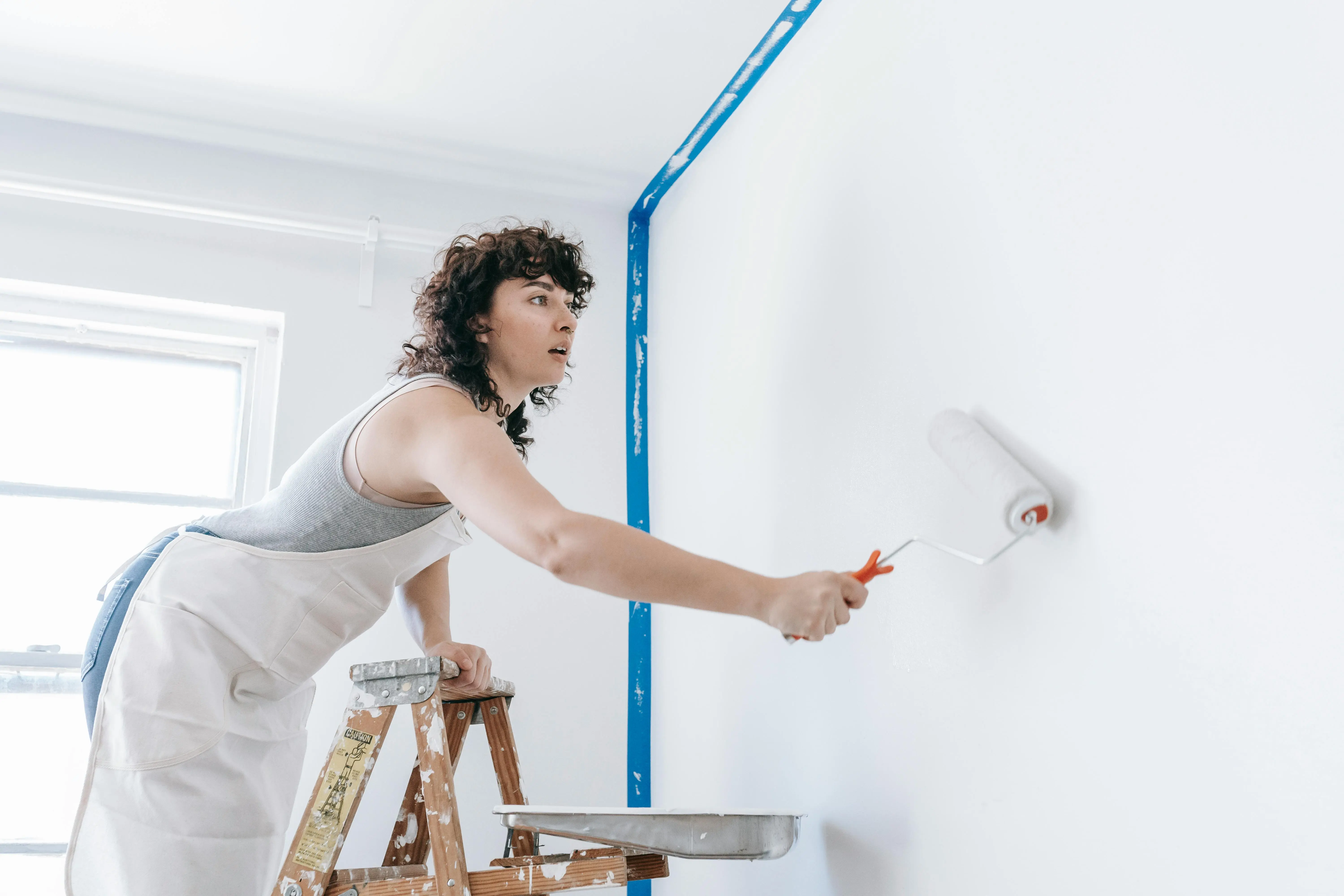 how much does a handyman charge to paint a room A woman stands on a ladder painting a wall white, part of a DIY home renovation.