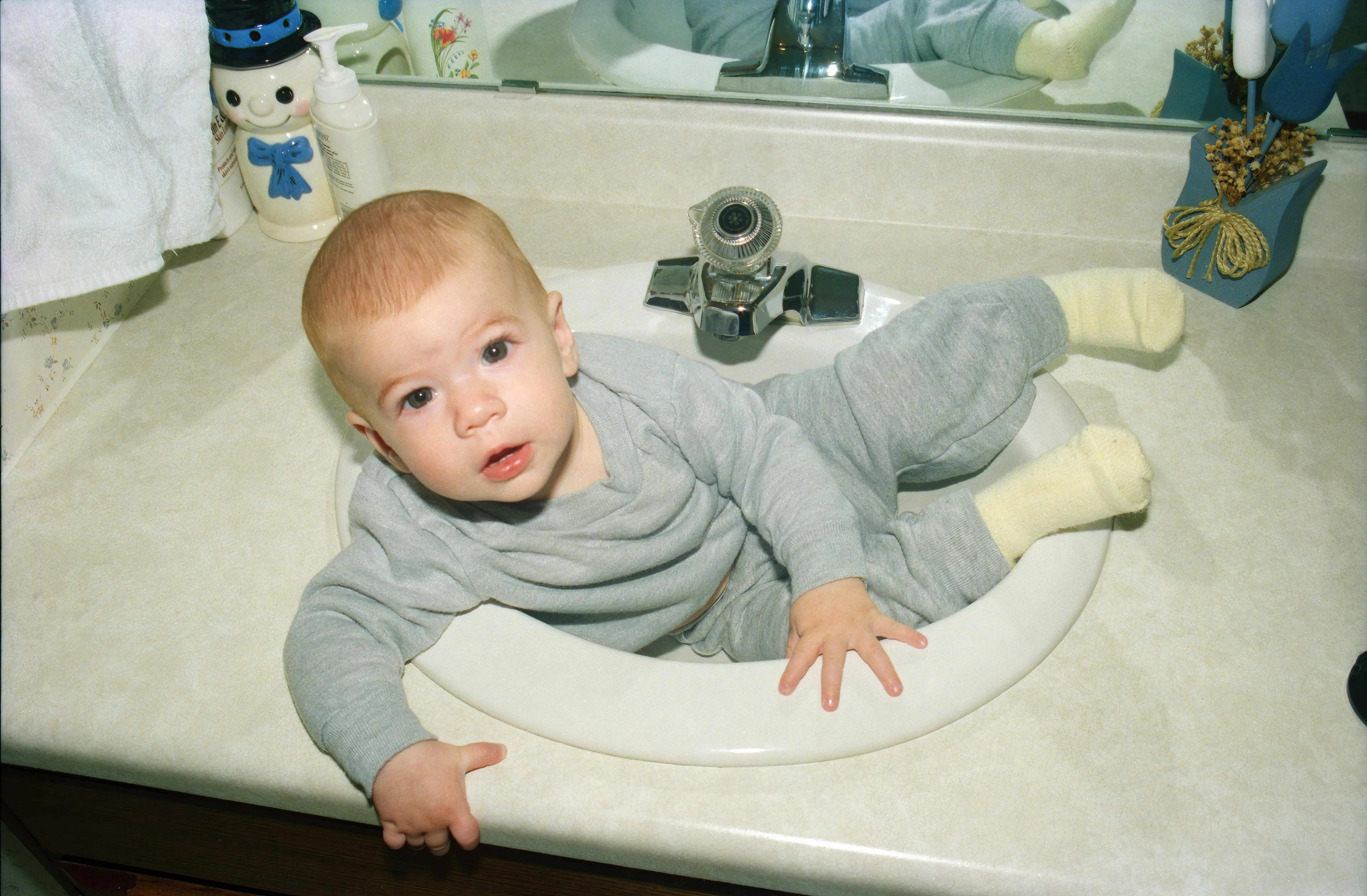 cracked sink repair Cute baby lying in a bathroom sink, wearing gray clothes, with mirror reflection.
