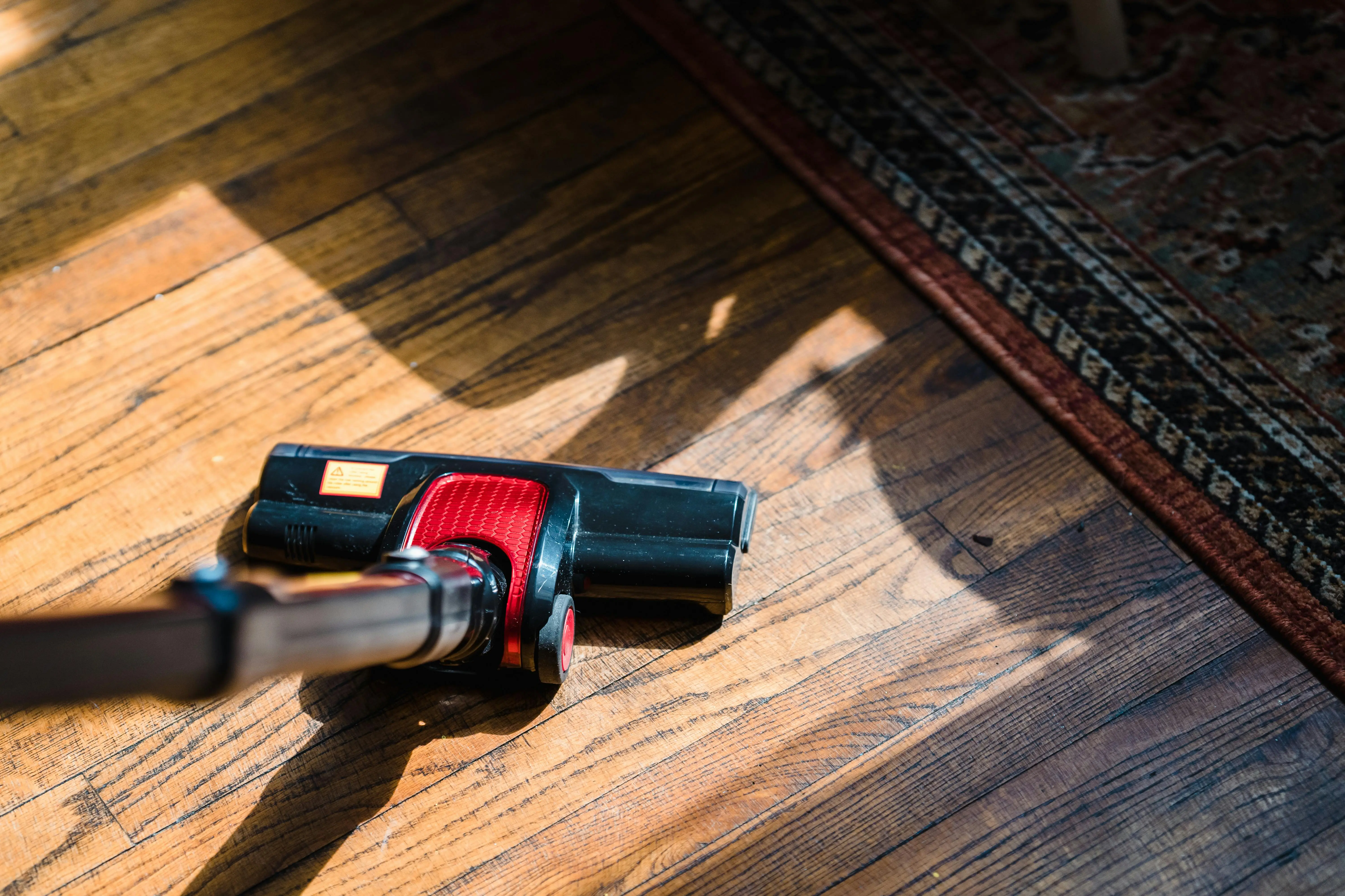 diy wood floor refinishing Close-up of a vacuum cleaner on wooden flooring near a patterned rug, showcasing domestic cleaning.