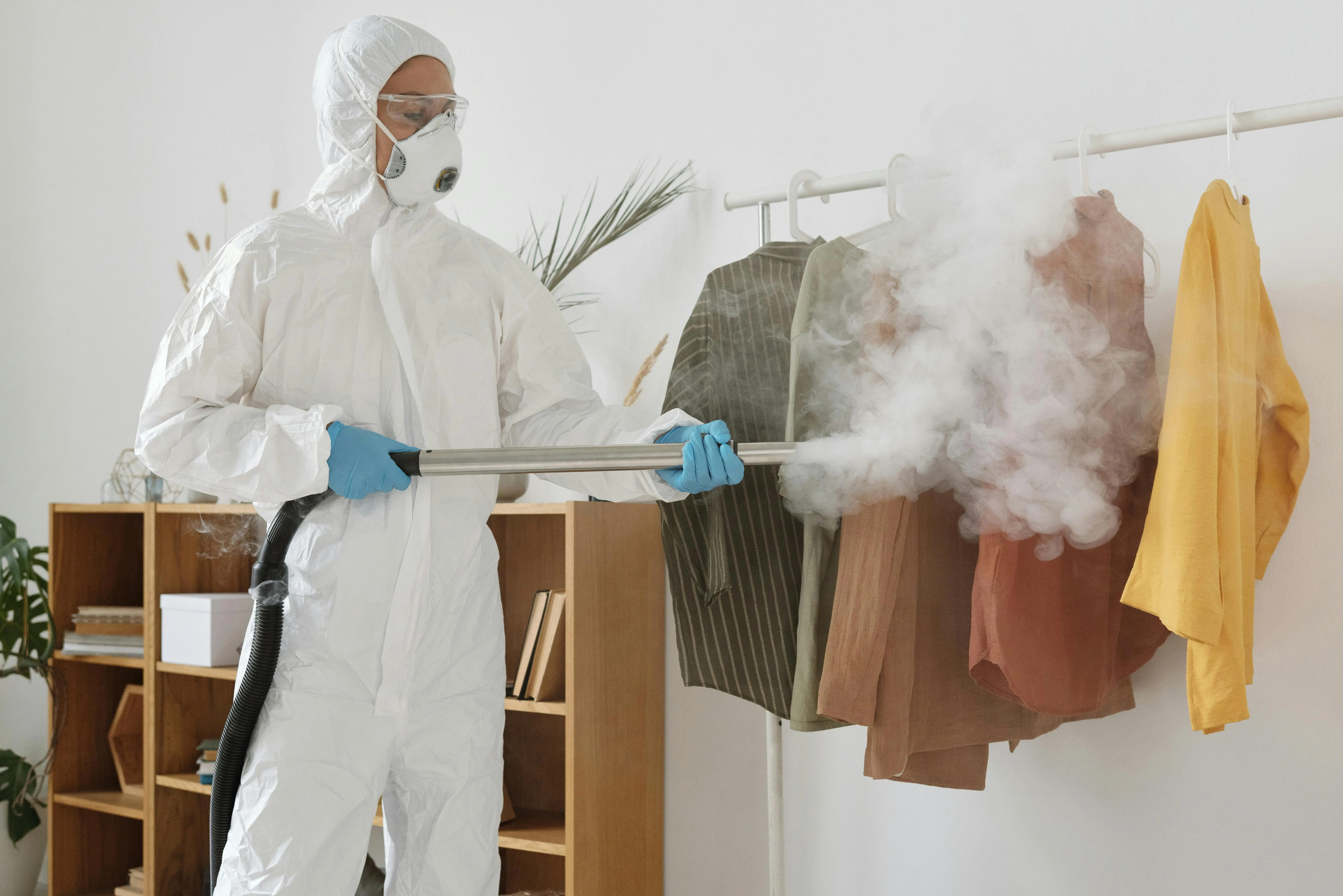 clean steam A person in PPE suit using a fumigation device to disinfect clothes indoors.