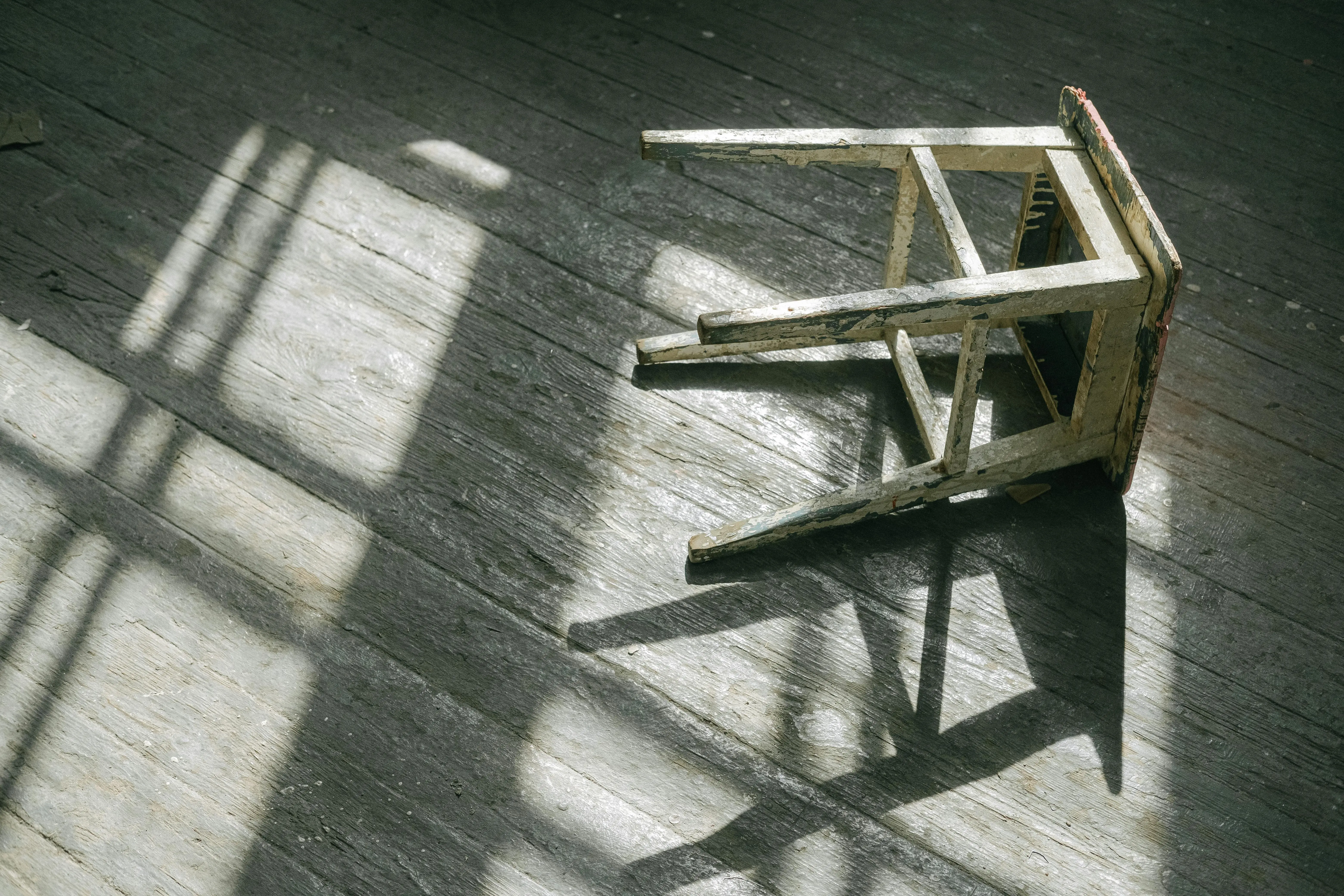 diy wood floor refinishing A worn wooden stool casting shadows on a sunlit wooden floor, creating a rustic and moody ambiance.