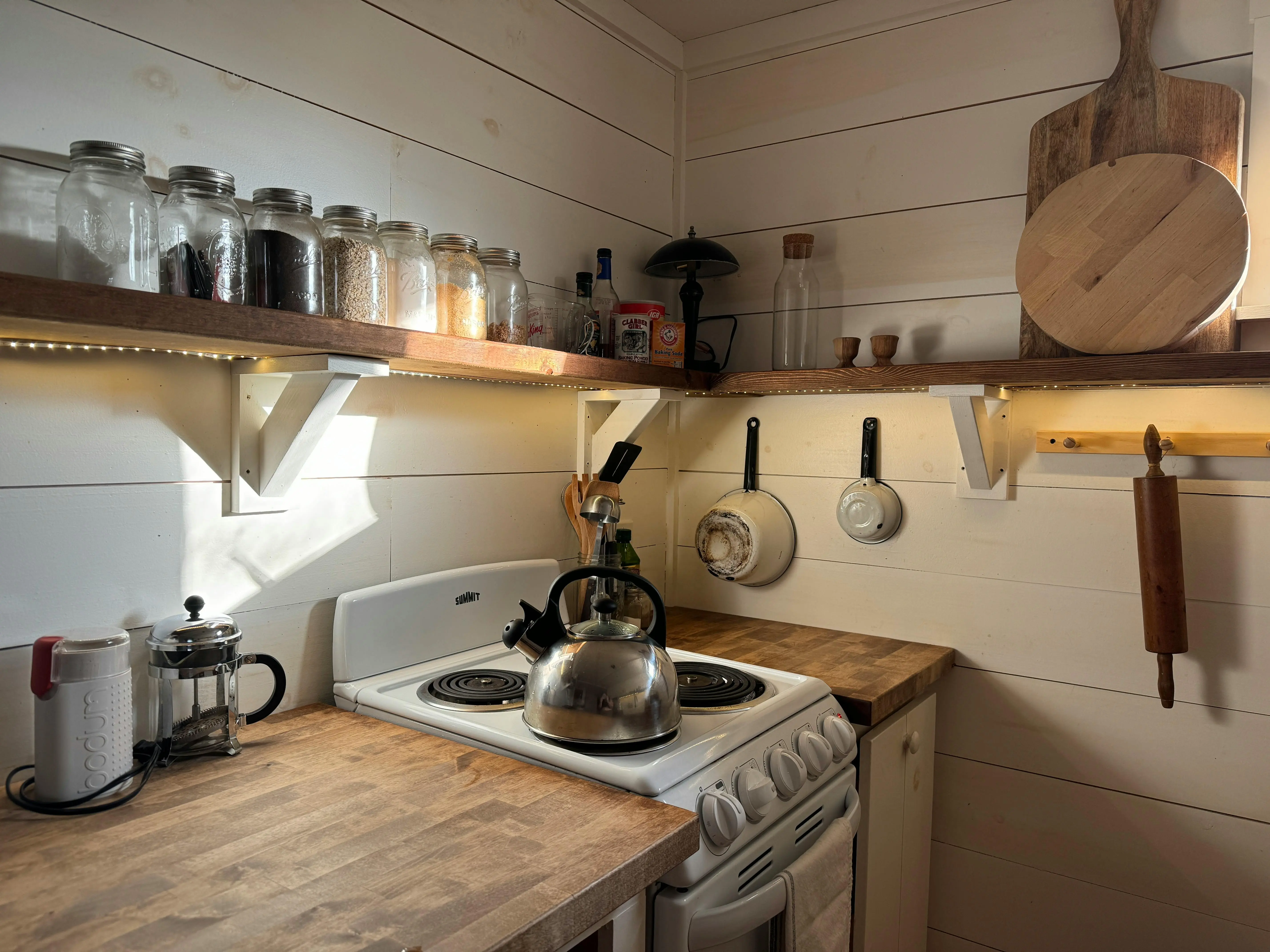 tiny home kitchen A small rustic kitchen with a wooden shelf, stove, and cooking utensils, offering a warm and cozy atmosphere.