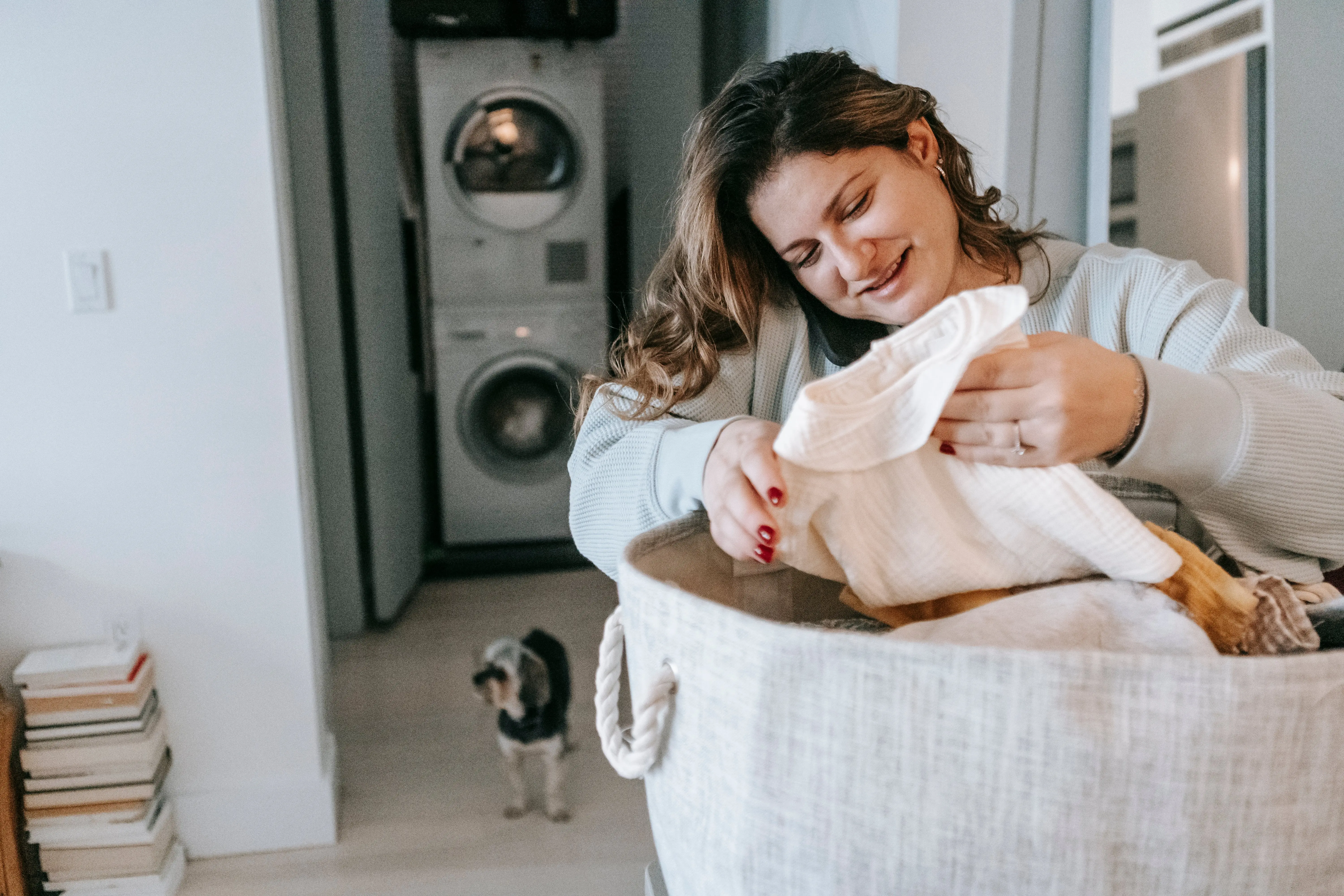 rolling laundry cart Optimistic female with clean clothes in basket having phone conversation while standing in light washroom with washing machines and dog
