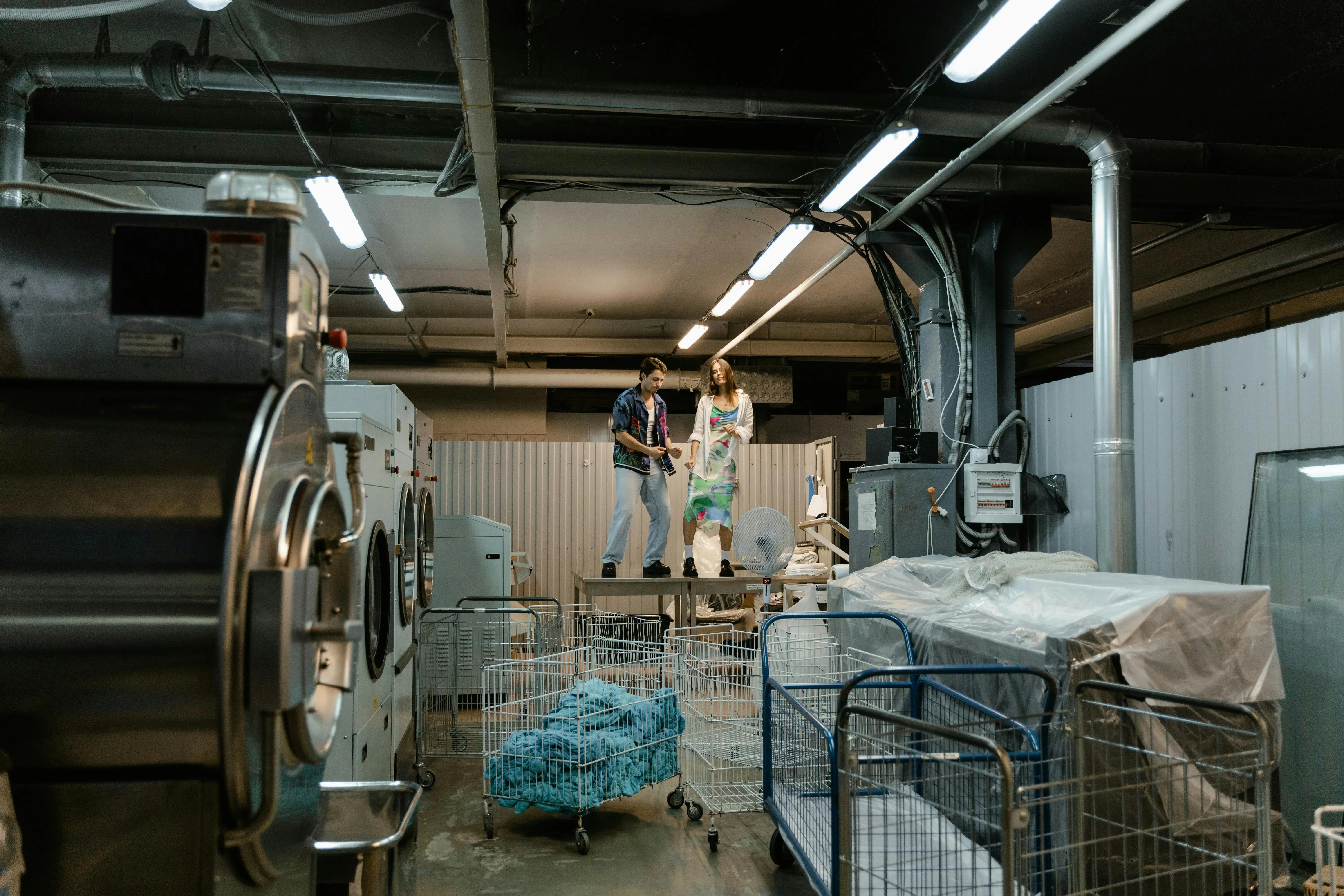 rolling laundry cart Two people working inside an industrial laundry facility with large machines and laundry carts.