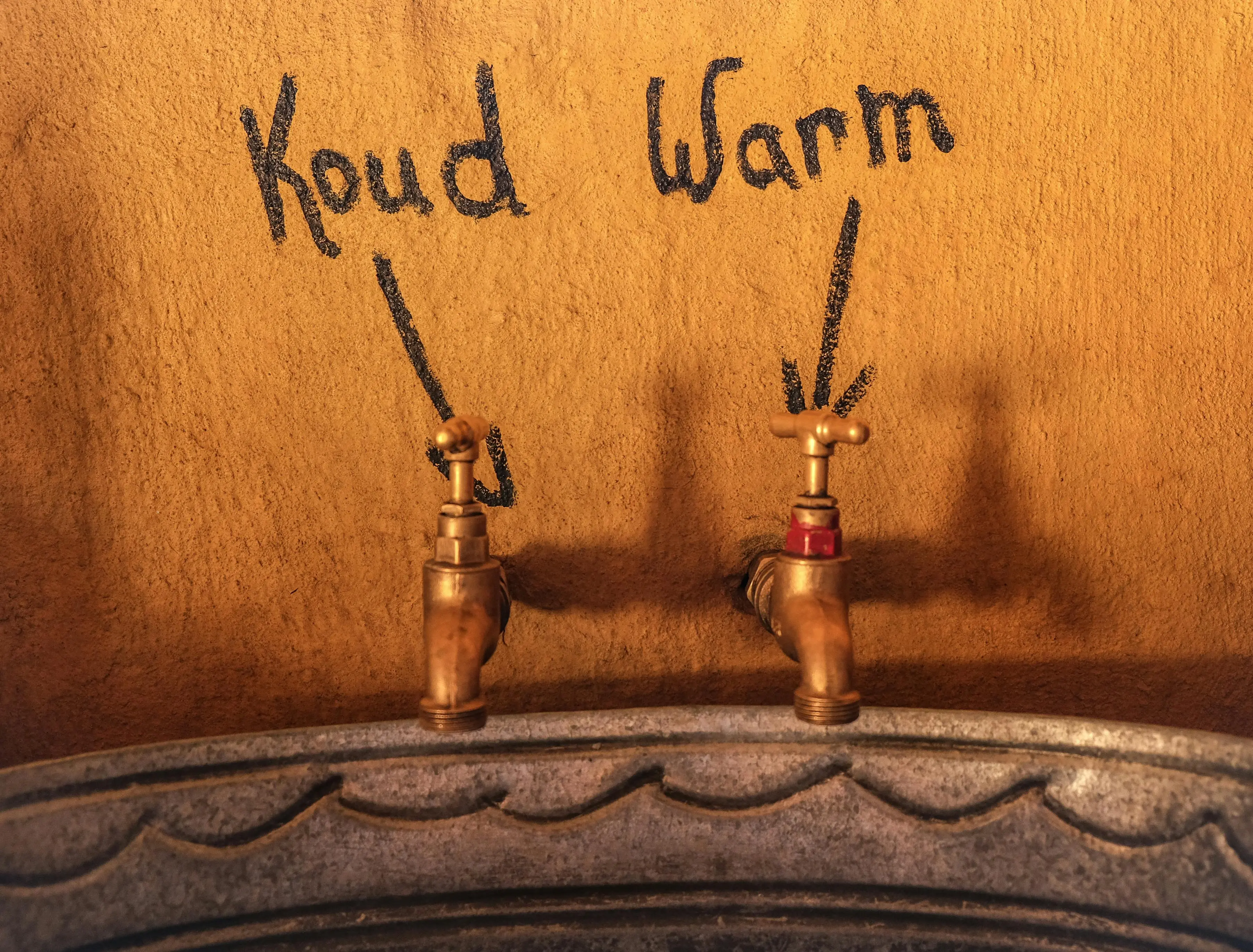 sink parts Close-up of vintage metal faucets labeled cold 'Koud' and warm 'Warm' on a textured gold wall.