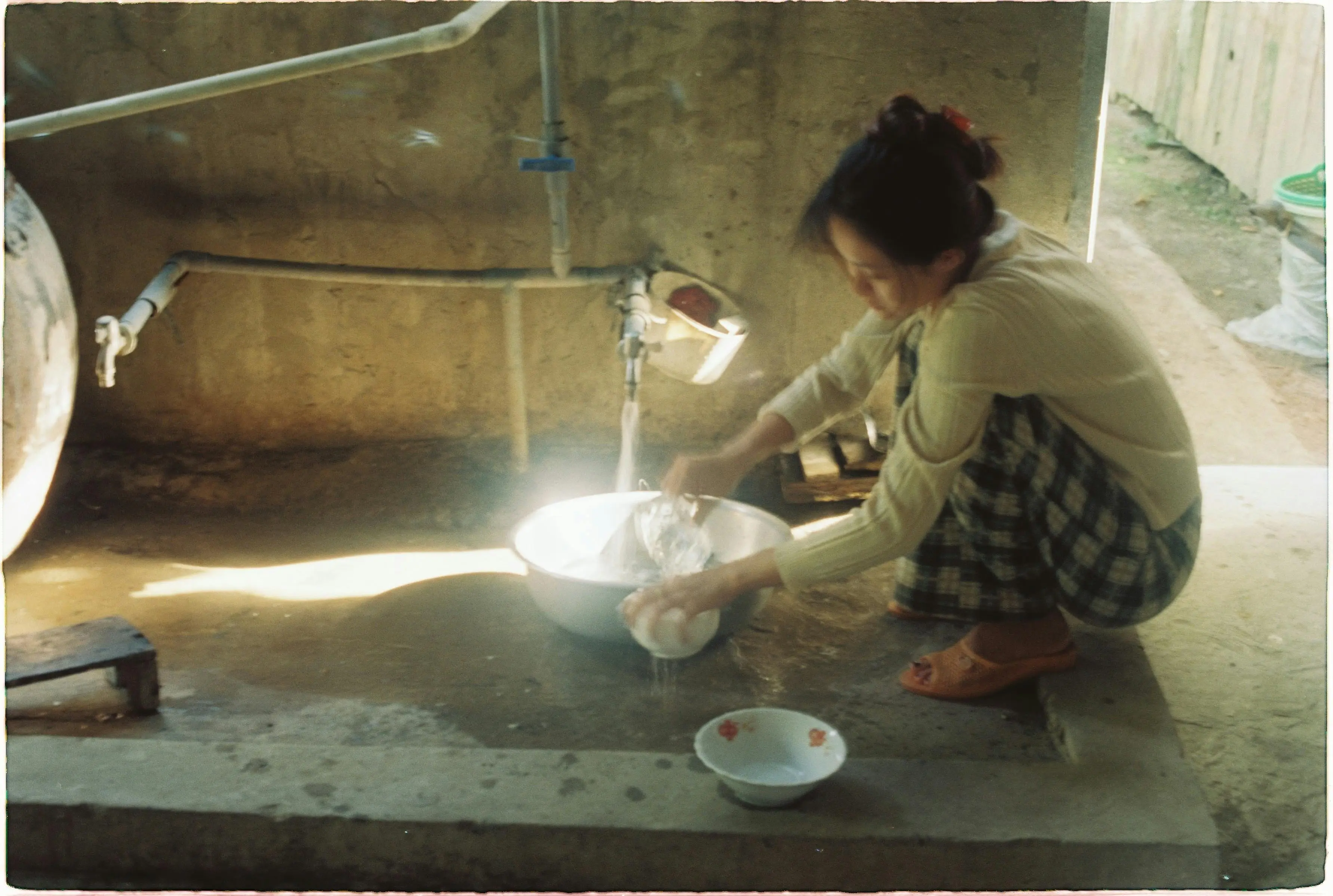 laundry signs A woman crouches while washing dishes in an outdoor sink with sunlight streaming in.
