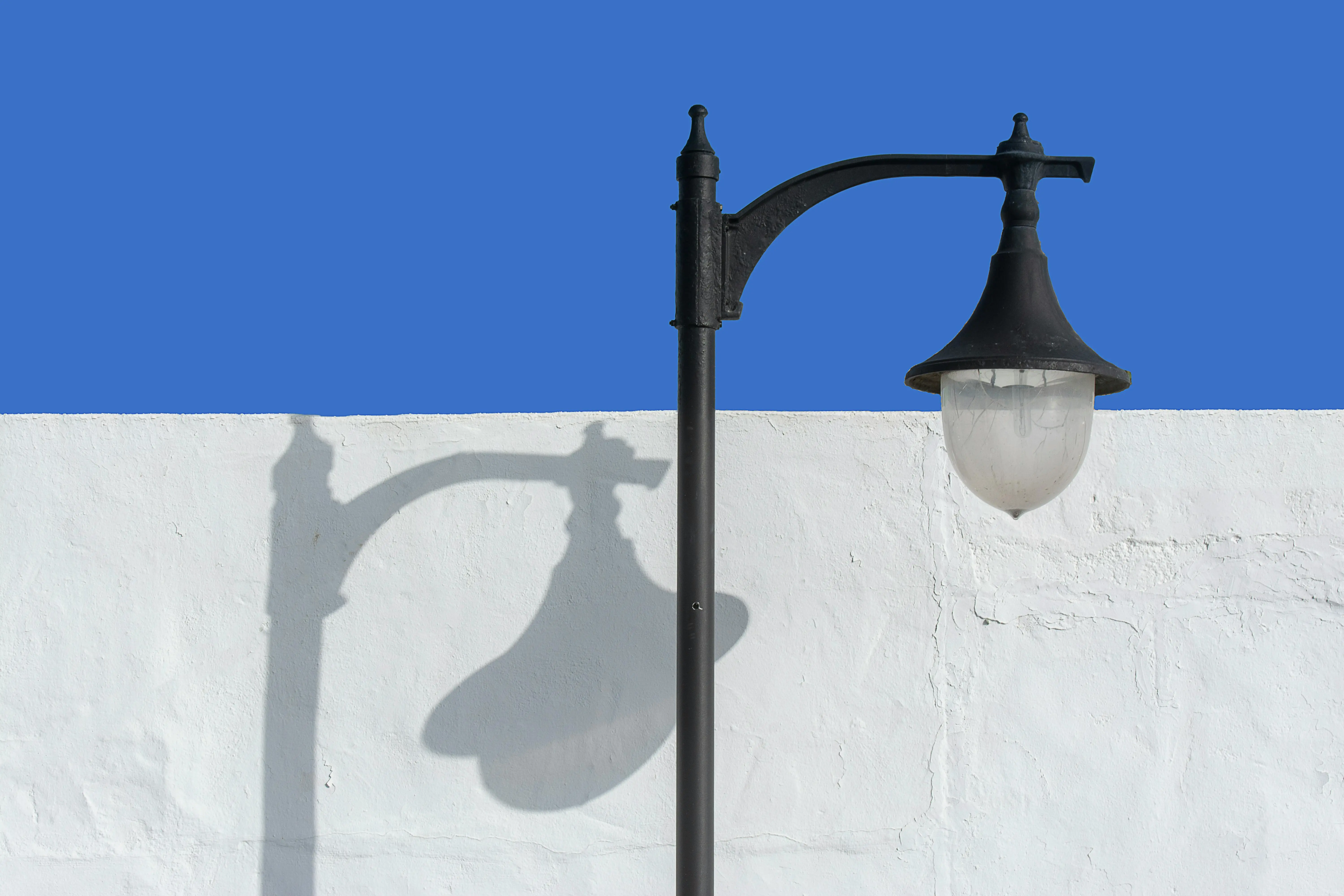 white lamp A vintage street lamp casting a shadow on a white wall under a clear blue sky.