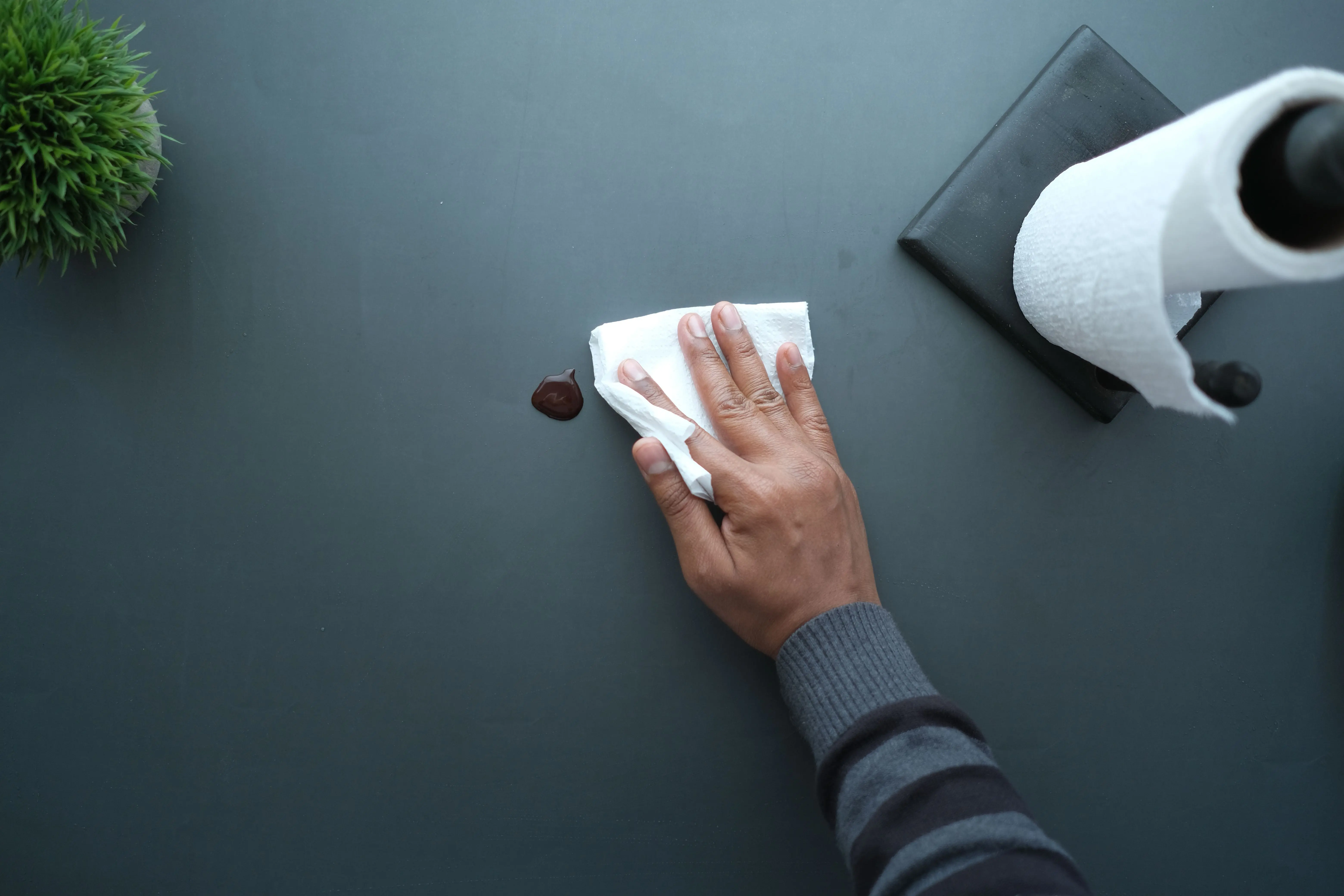 and paper towels A hand wipes a dark surface with a tissue near a paper roll and plant.