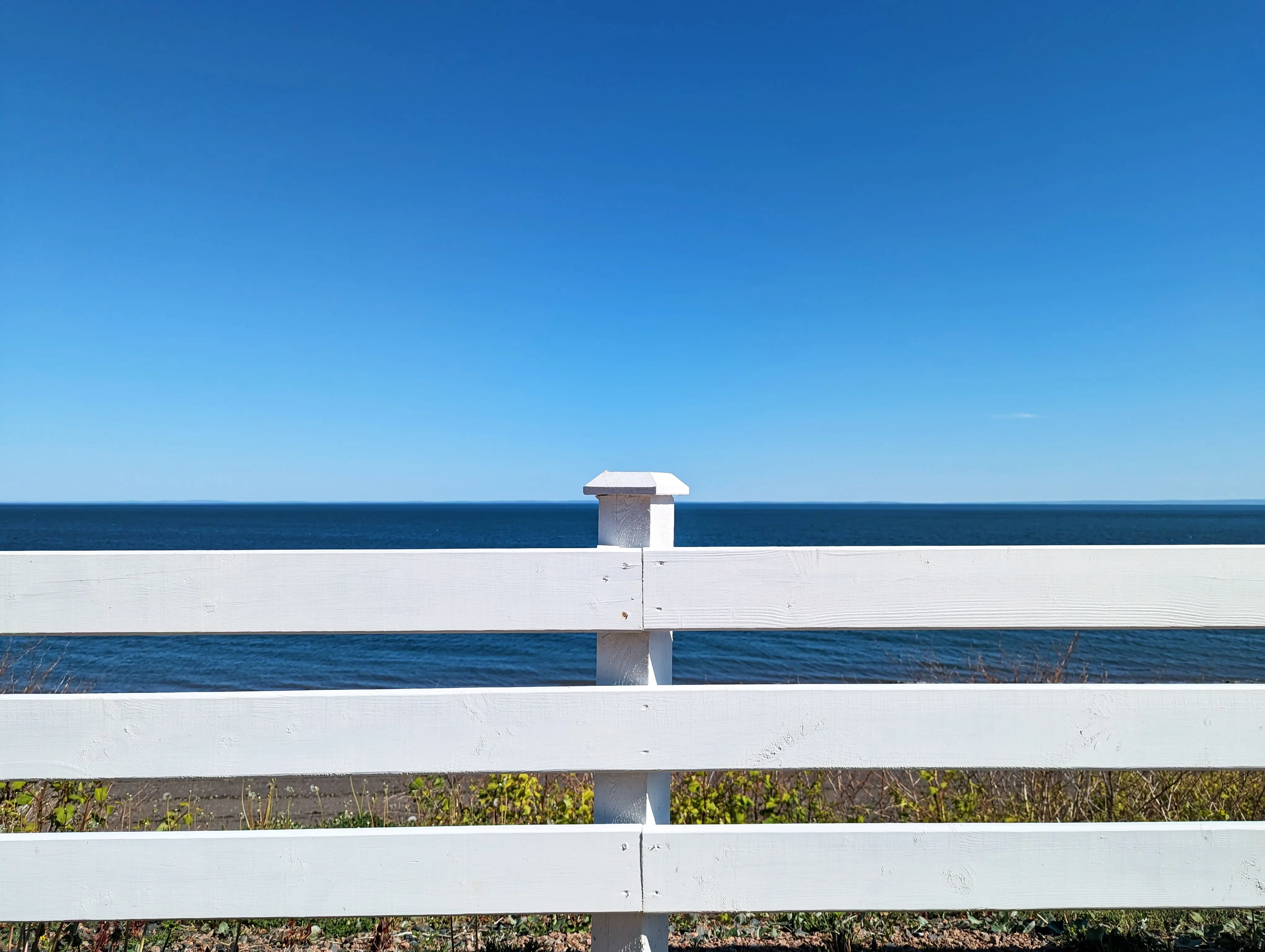 vinyl fence parts Serene view of a white wooden fence facing the ocean under a clear blue sky in summer.