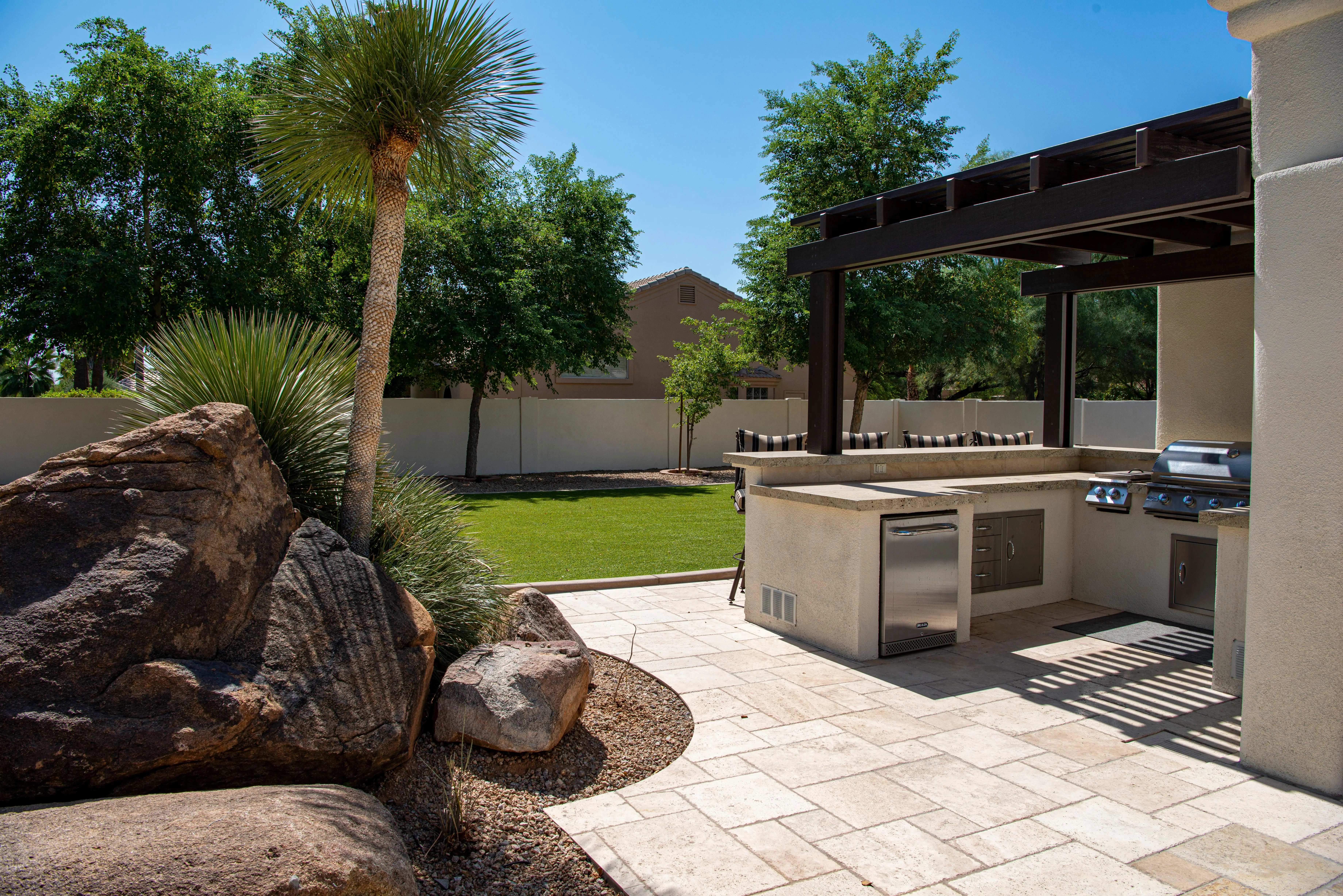 paver patterns Stylish outdoor kitchen on a sunny patio with lush landscaping and clear blue sky.