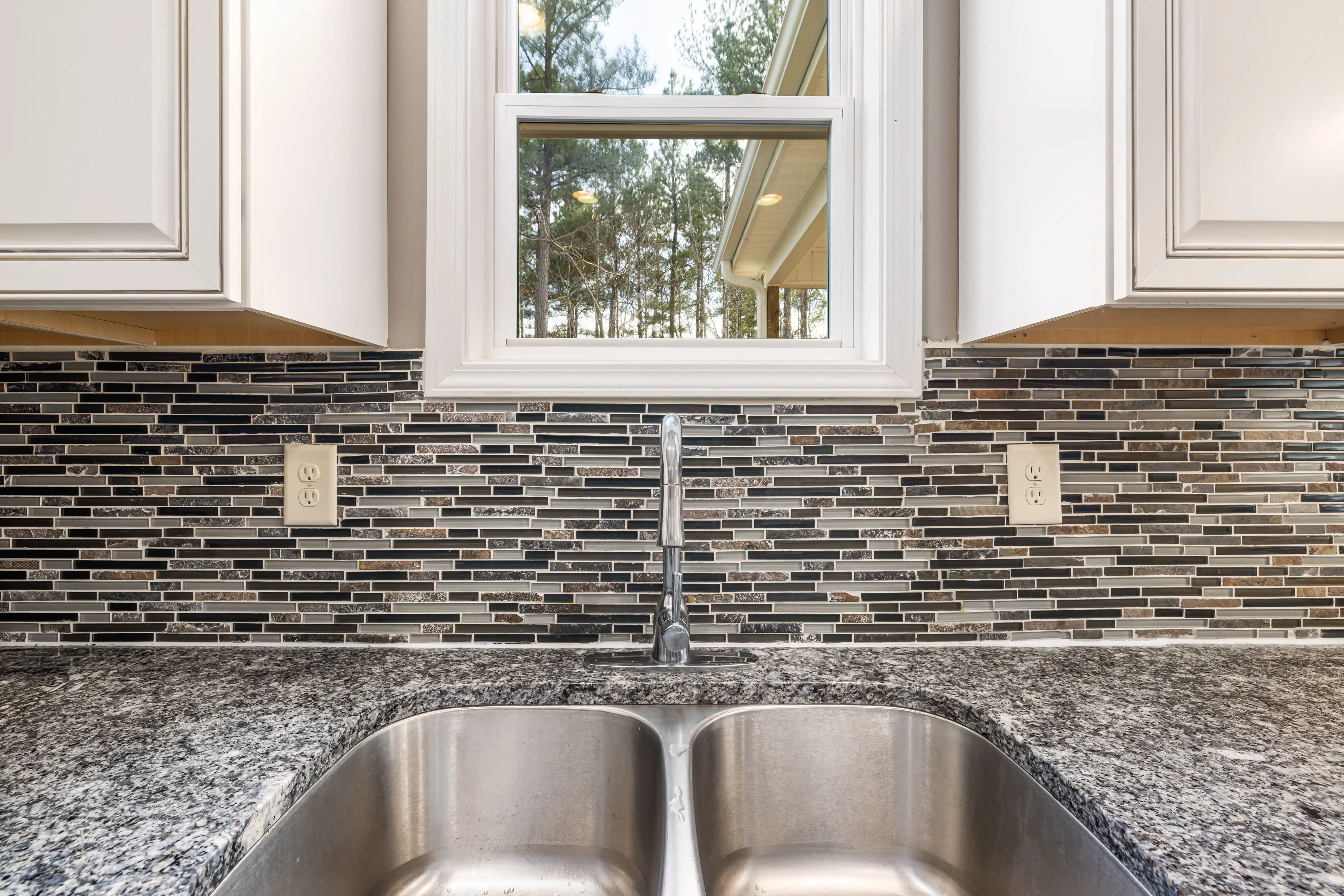 modern kitchen backsplash Close-up of a stainless steel sink on a granite countertop with a tiled backsplash and a window view.