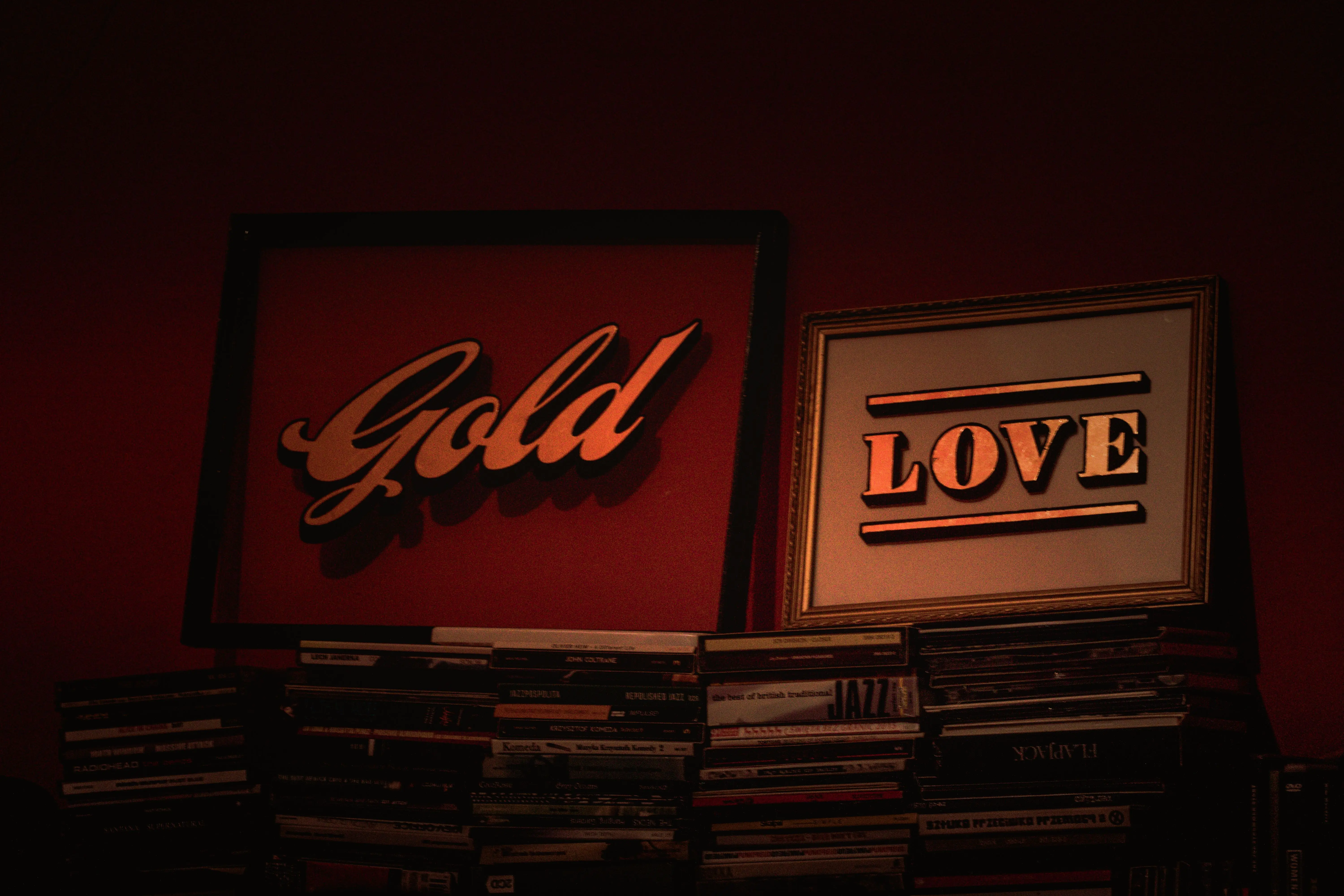 gold wall art Artistic display of framed 'Gold' and 'Love' signs above stacks of CDs on a dark red wall.