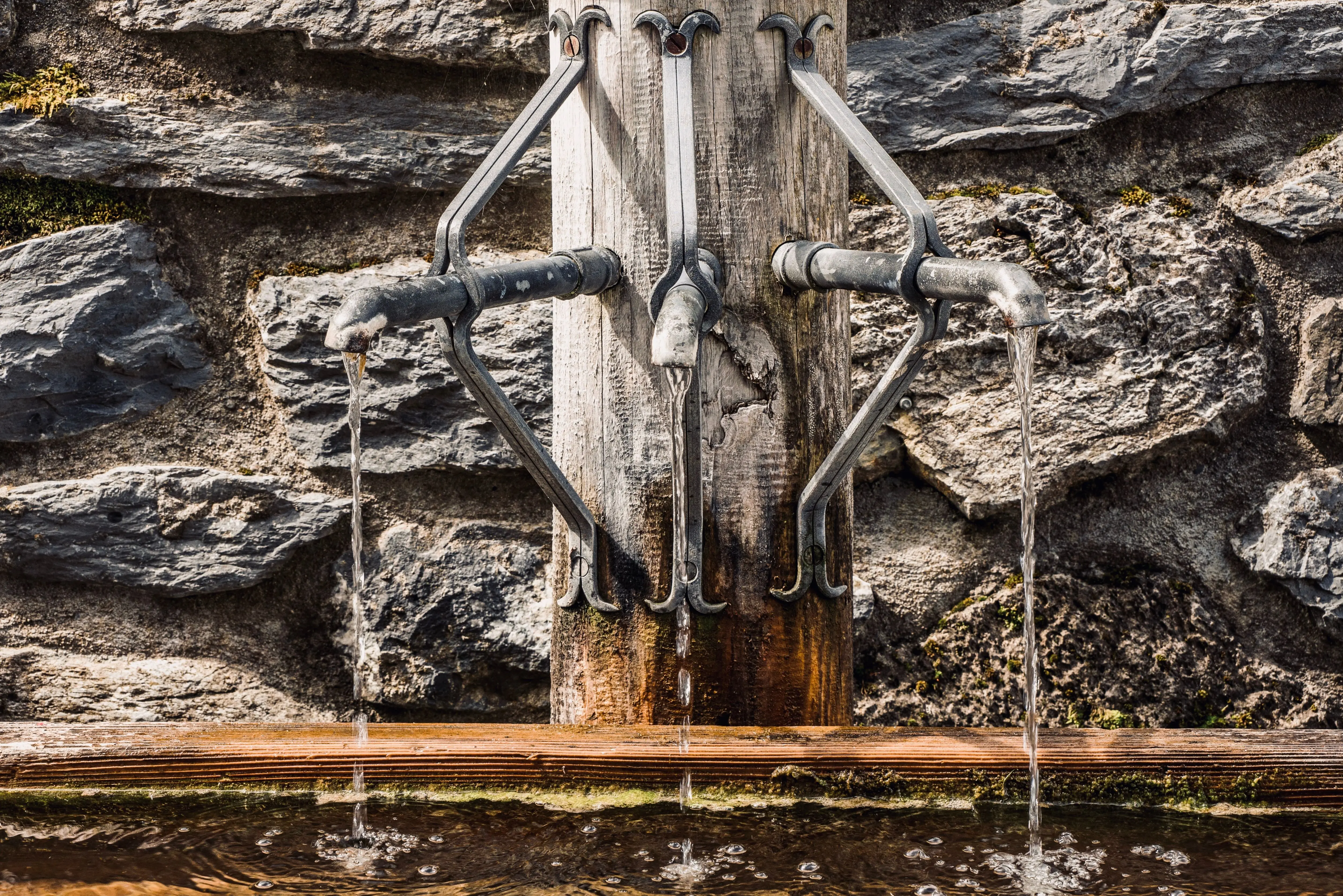 outdoor wall decorations metal Metal taps of fountain with pouring water into tank placed on wooden post near stone wall on street in city