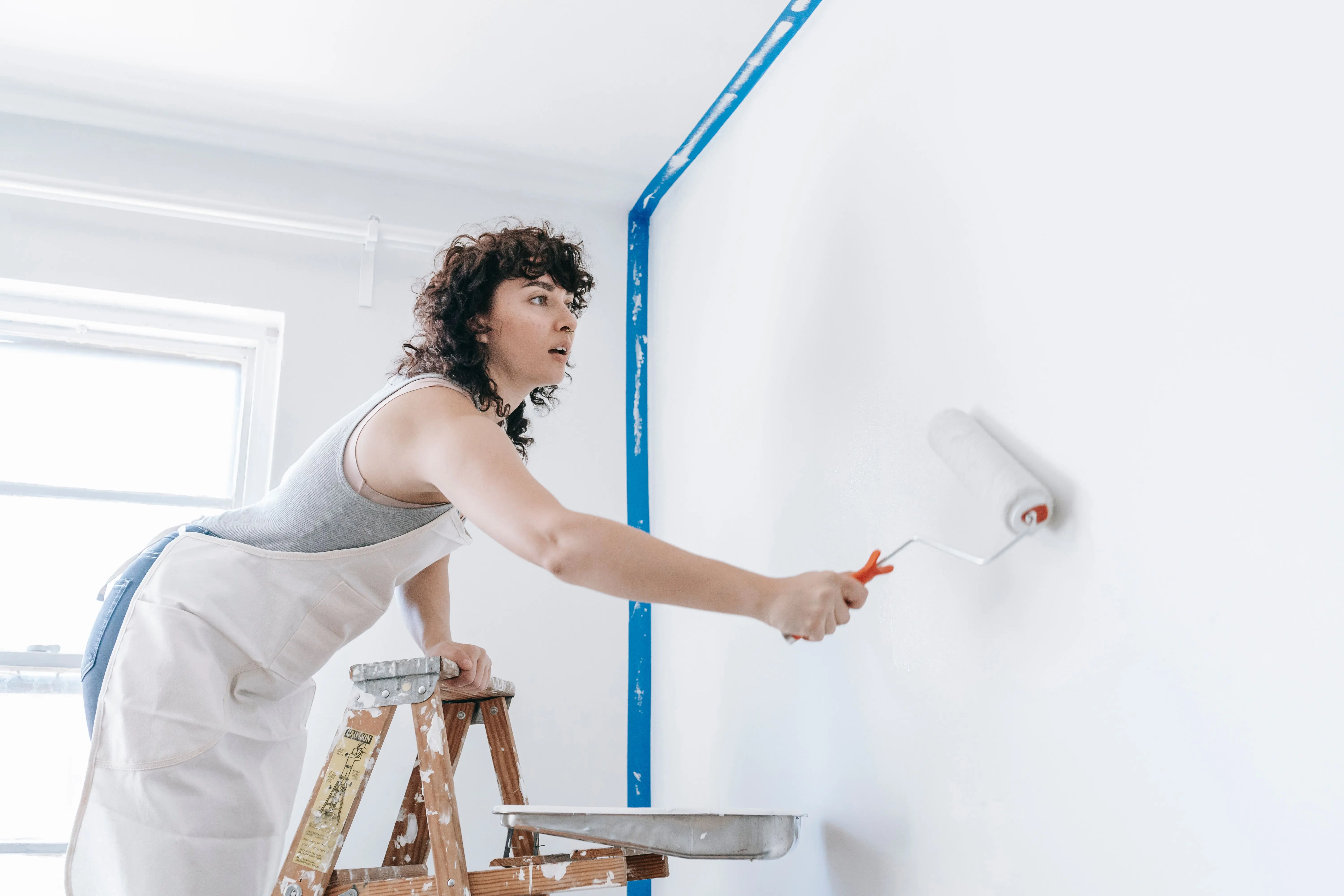 painting price per square foot A woman stands on a ladder painting a wall white, part of a DIY home renovation.