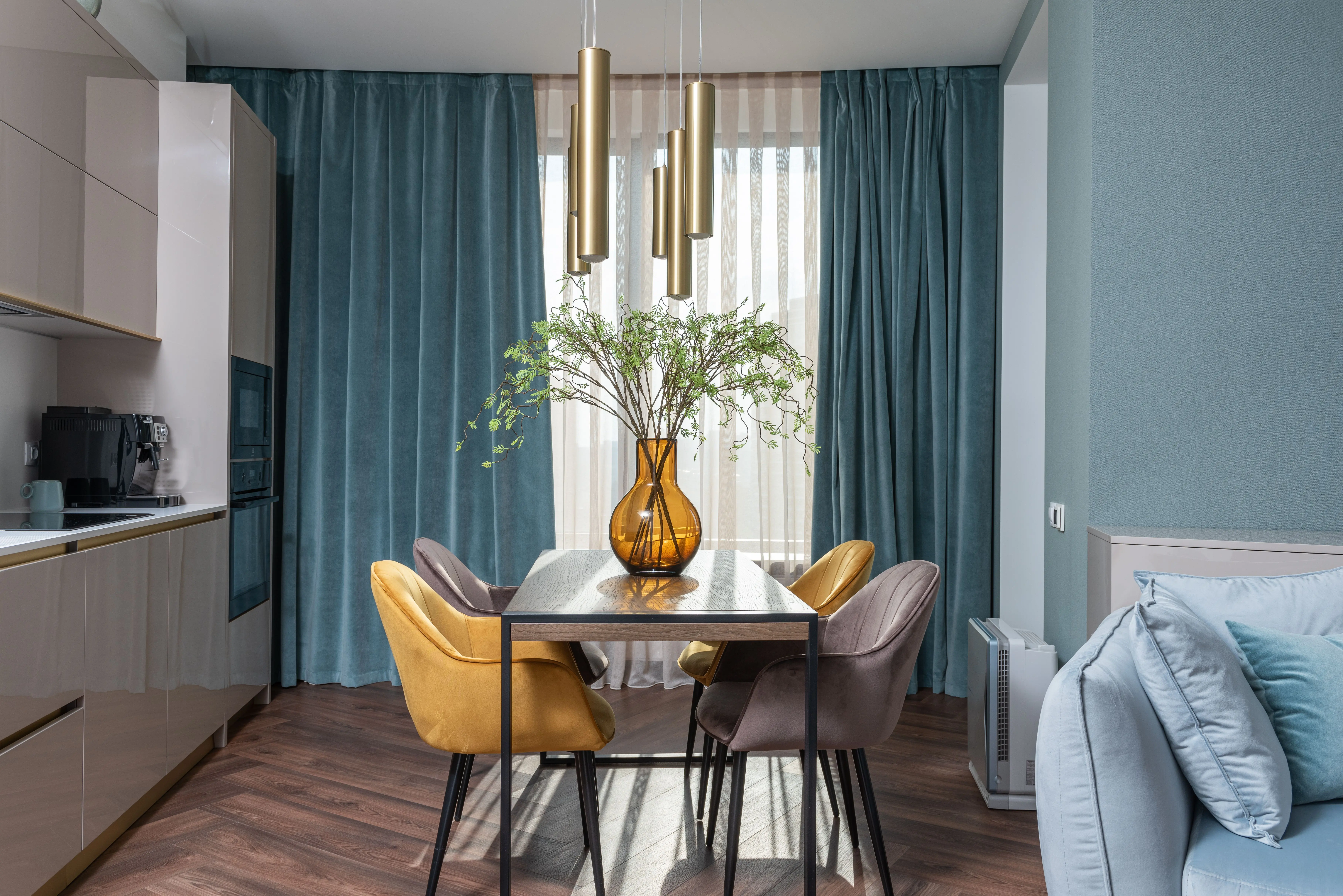 patterned drapes Glass vase with flowers on table with chairs placed near kitchen cabinets and soft couch in modern apartment with window