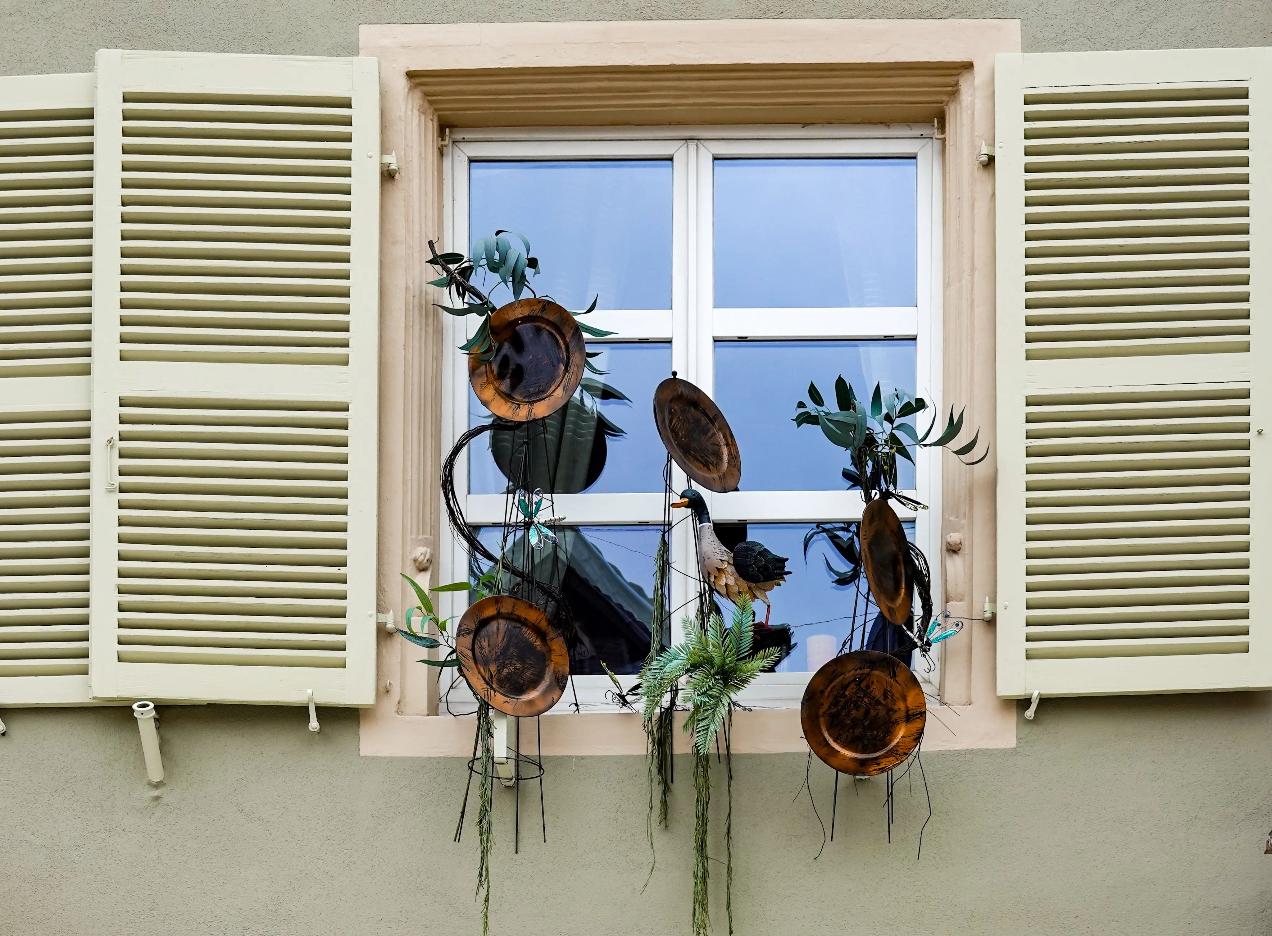 outdoor wall decorations metal A quaint window with shutters adorned by metal art and greenery, reflecting a cozy old town charm.