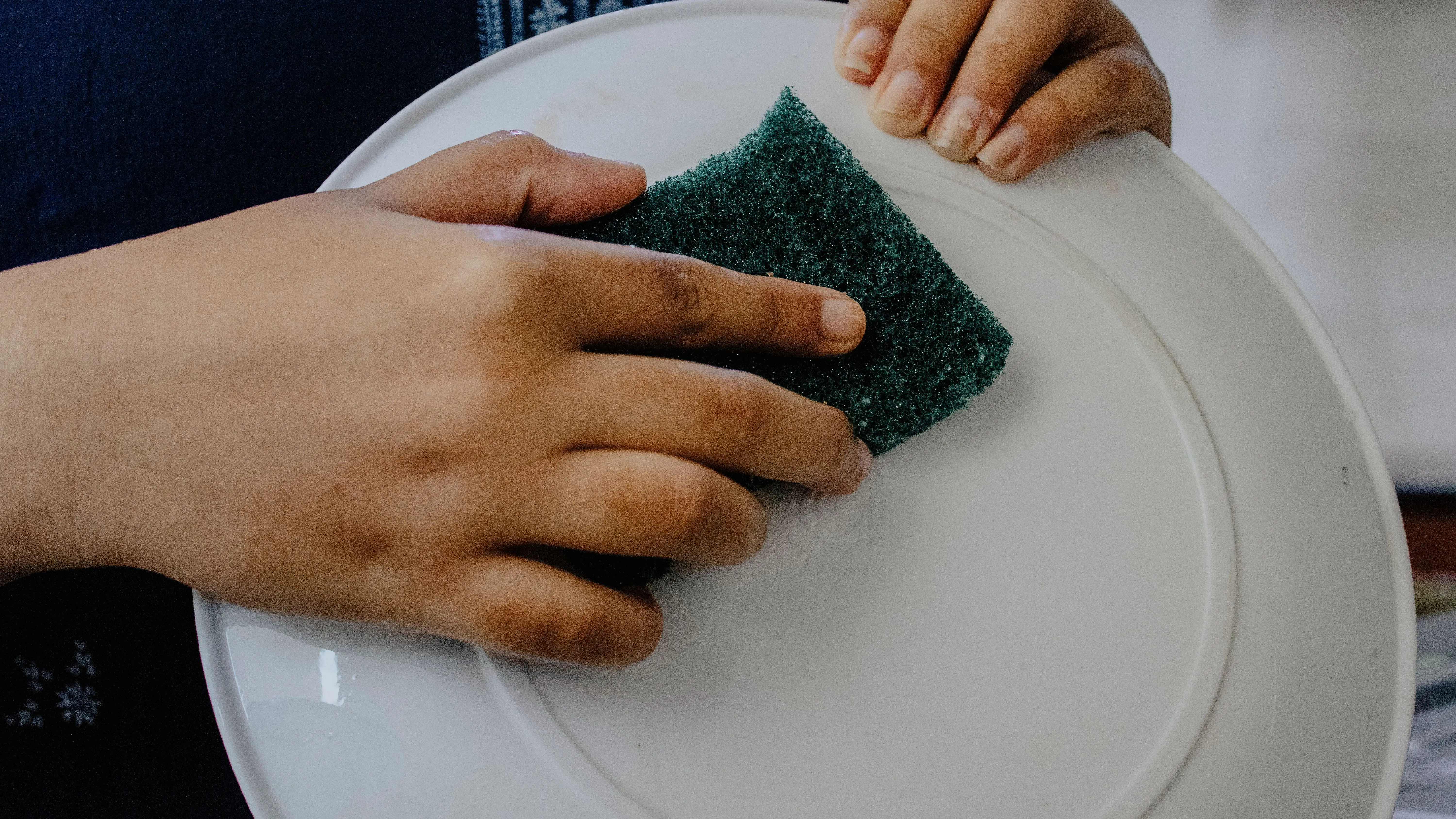 how to get grass stains out Hands using a green scrub pad to clean a white plate indoors.