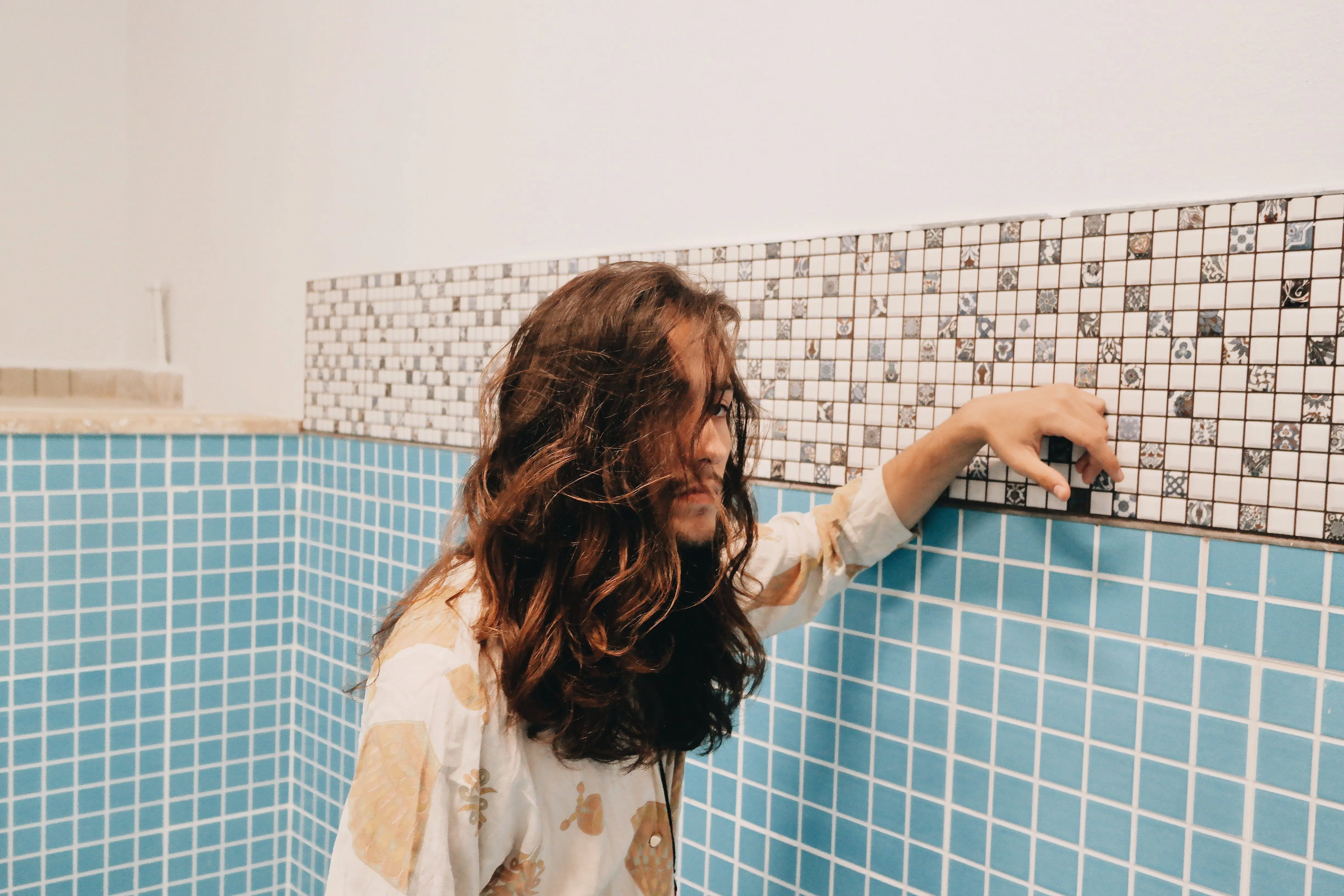 blue subway tile Young man with long hair posing indoors against a decorative tiled wall.
