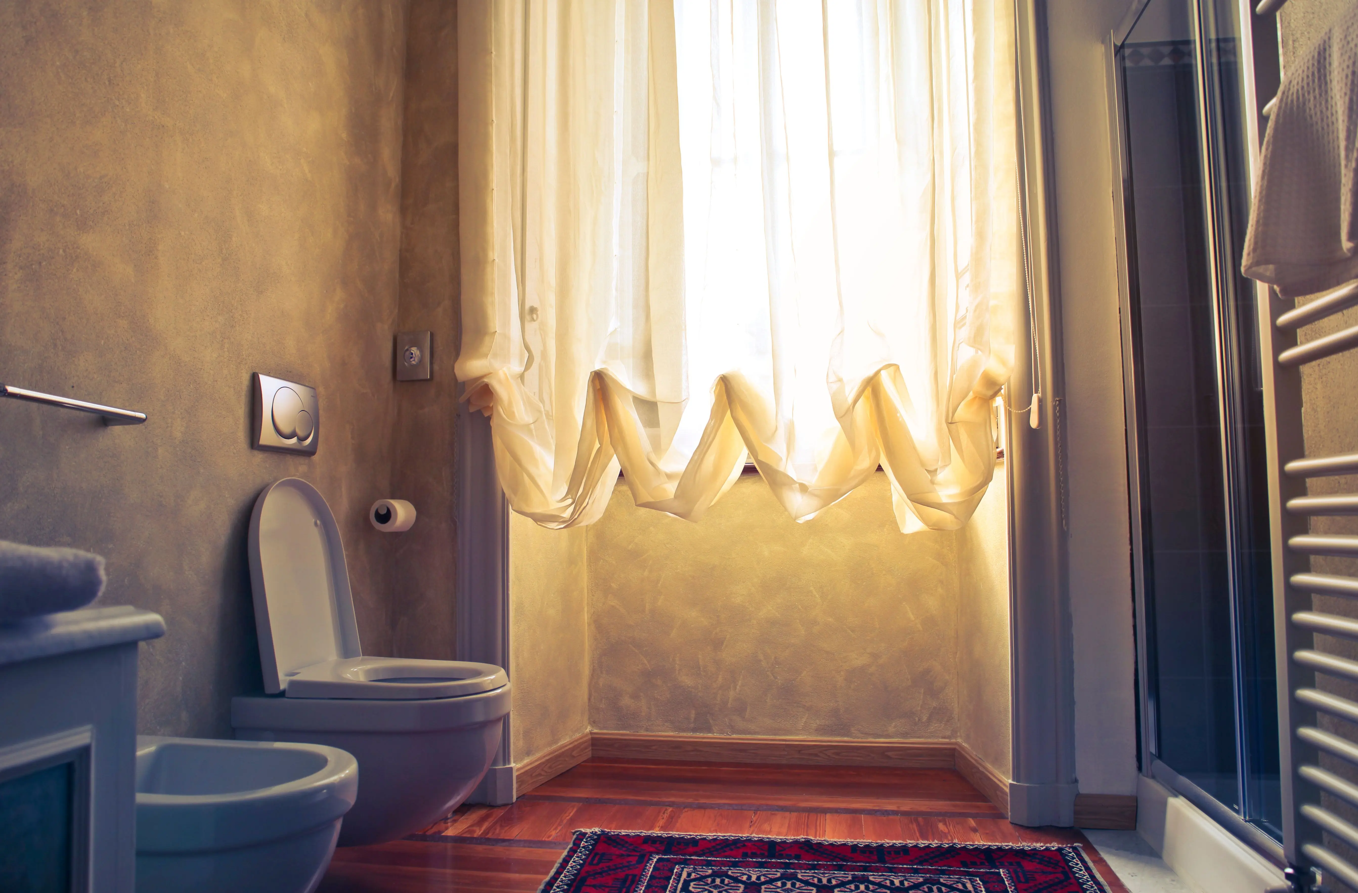 blue floor tile Warmly lit bathroom with a bidet, toilet, and a flowing curtain revealing soft sunlight.